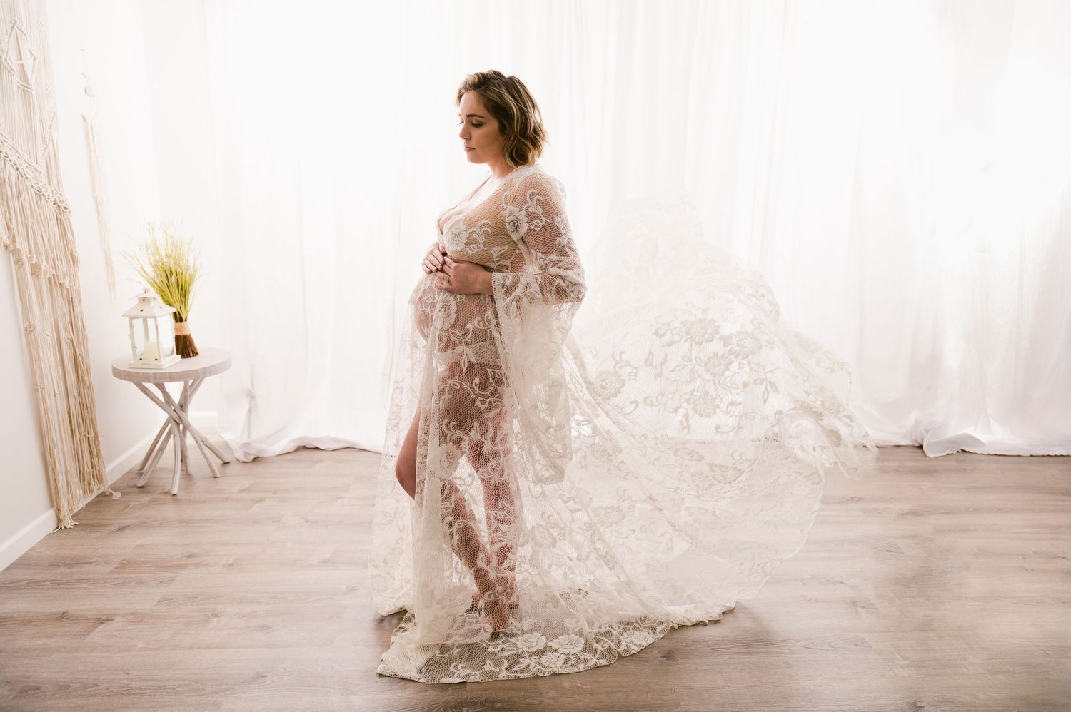 Woman in a delicate lace maternity gown poses in light-filled studio with white curtains and wooden floor.