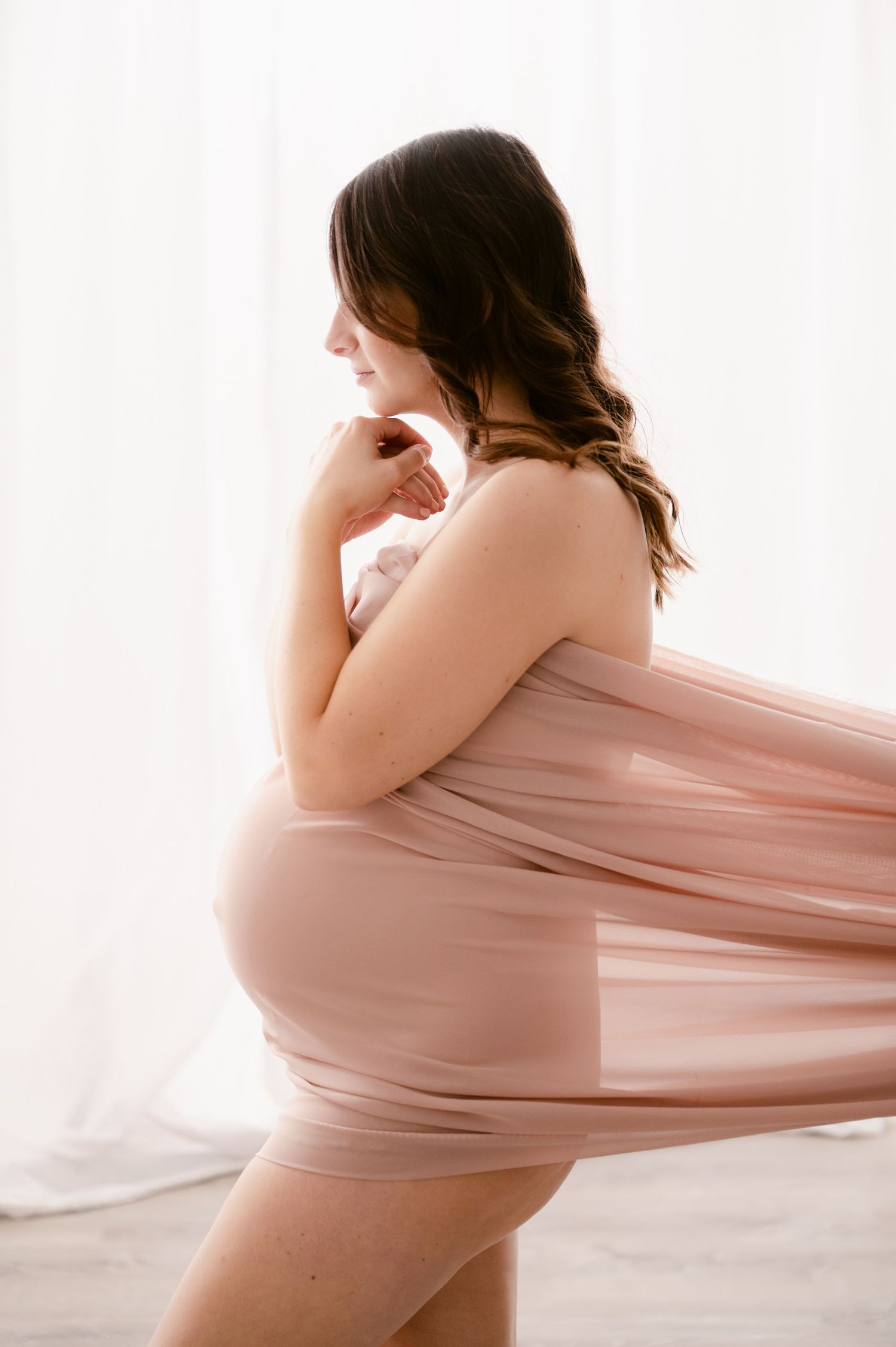 Artistic maternity photo featuring flowing pink fabric draping over a pregnant silhouette against white backdrop.