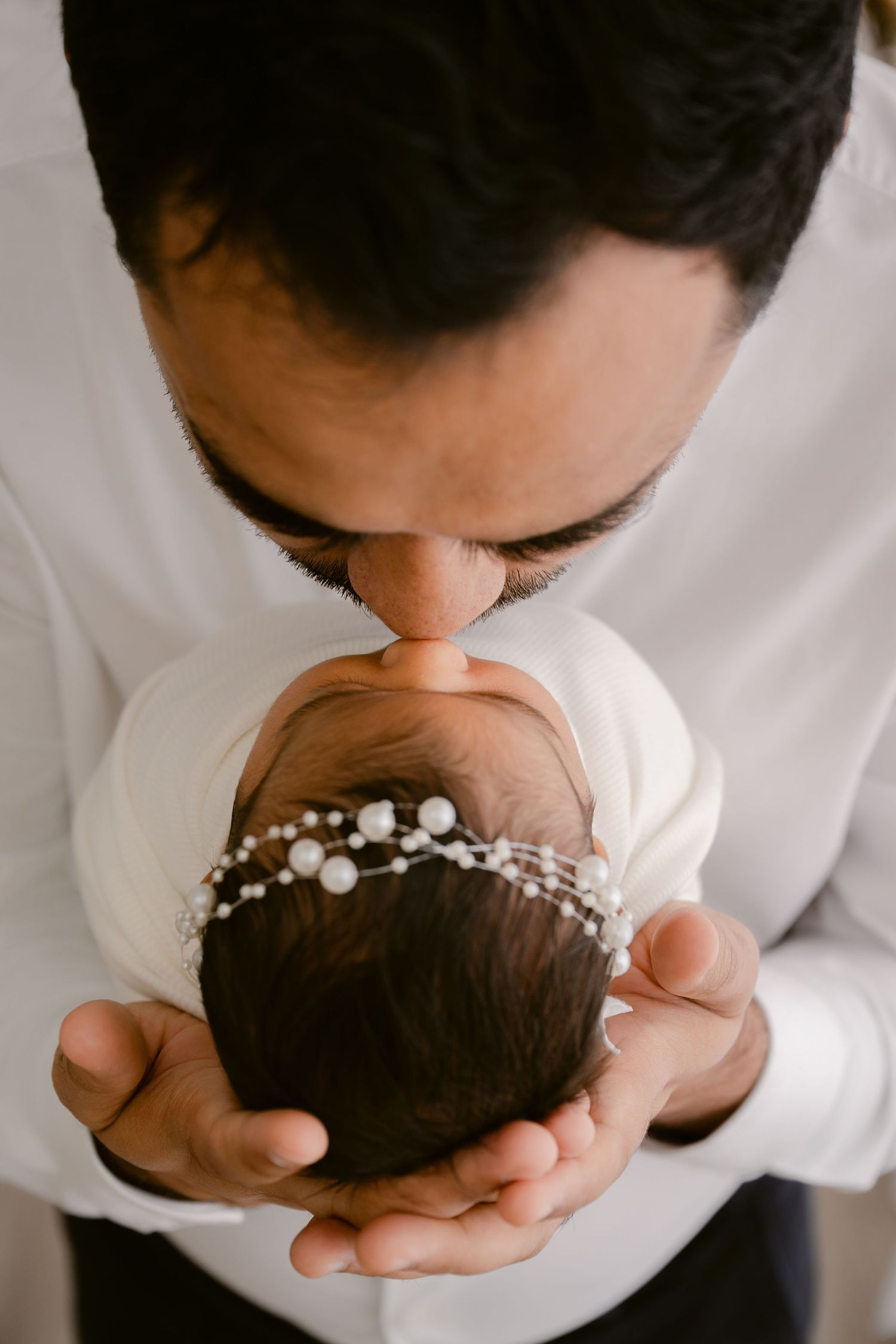 Parent kissing newborn baby's head adorned with delicate pearl headpiece.