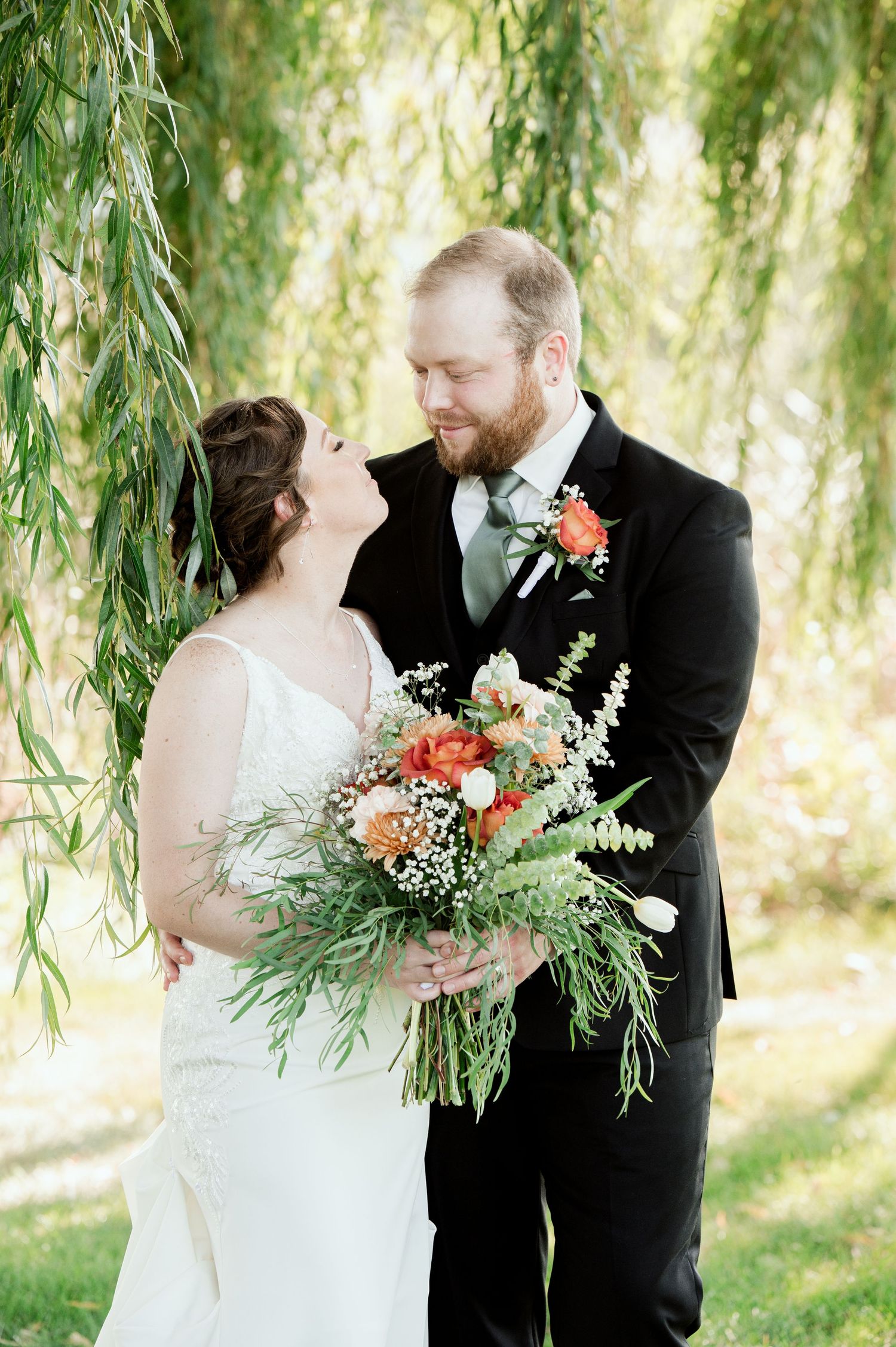 The Grands At Mulligans. A couple shares intimate moments under hanging willow branches at their outdoor wedding ceremony.