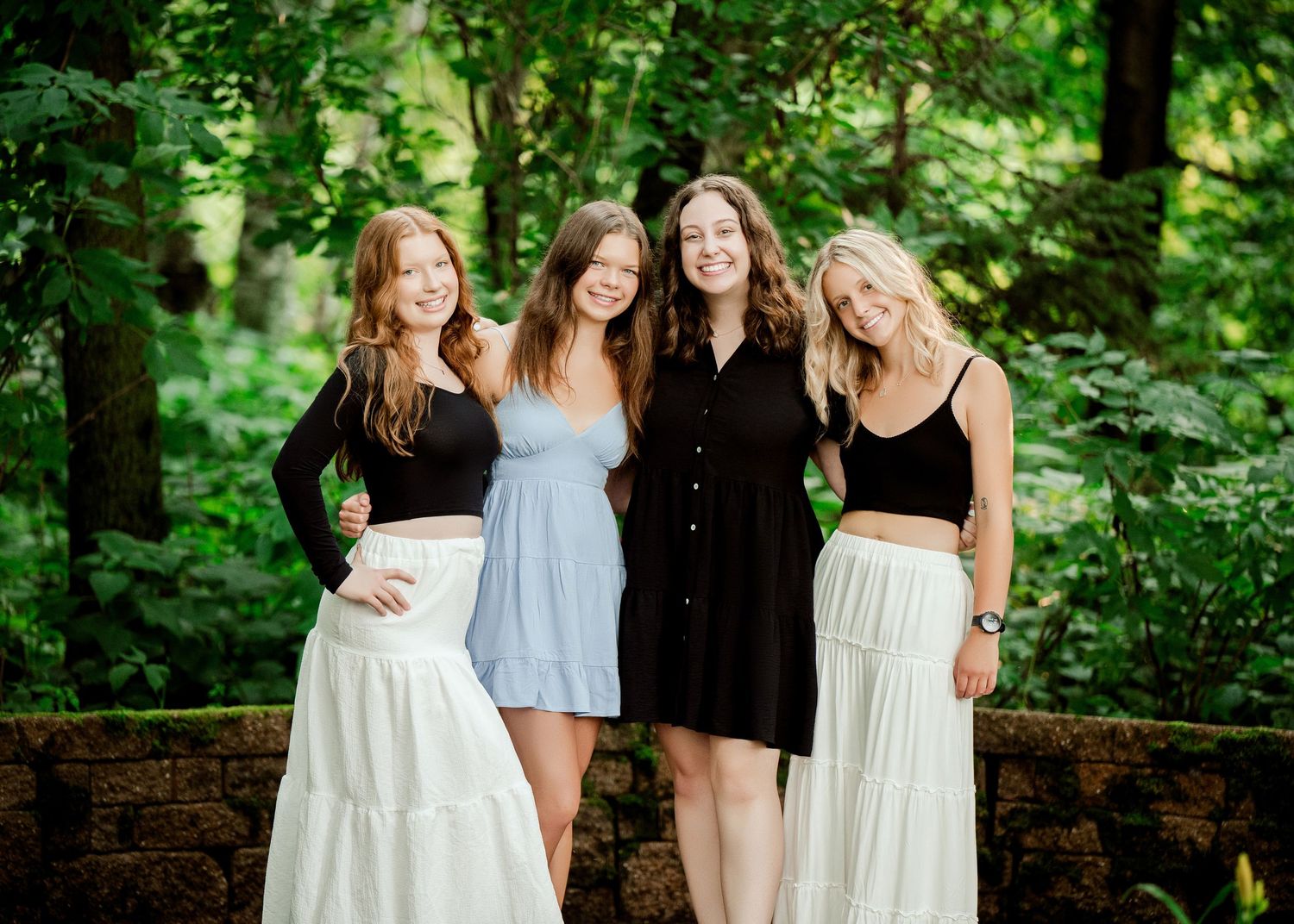 Clear Lake MN Family Photos:Four people in coordinated black and white outfits pose together in front of lush green foliage on a summer day.
