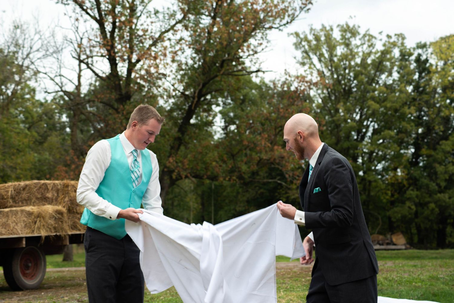 A couple in formal attire playfully tugs at opposite ends of a white sheet in a grassy outdoor setting.
