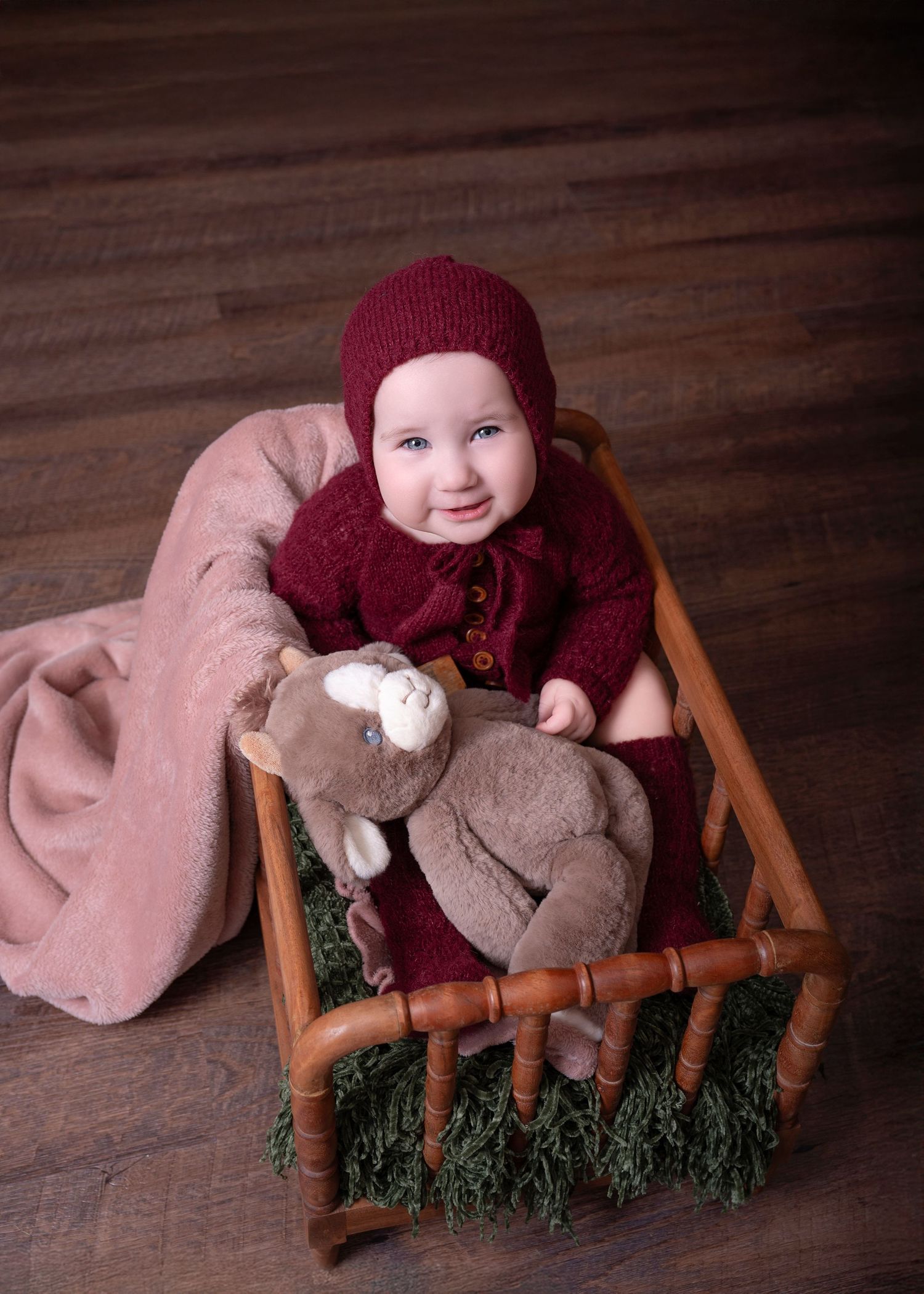 Baby in burgundy knit outfit sitting in wooden chair with plush toy.