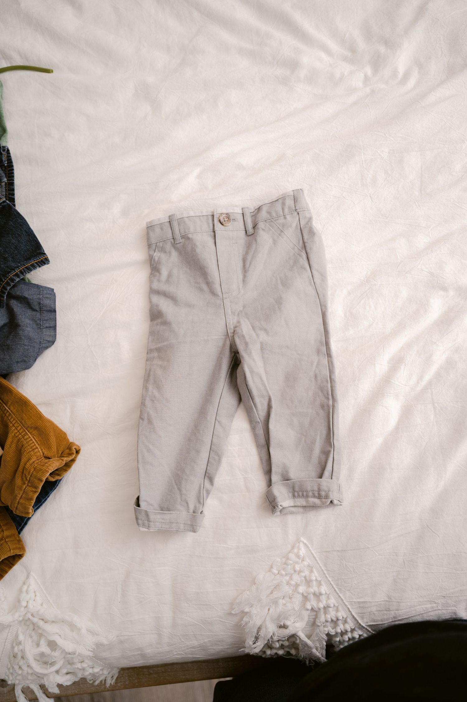 Light gray toddler pants with rolled cuffs lay flat on white bedding with partial views of other clothing items visible.