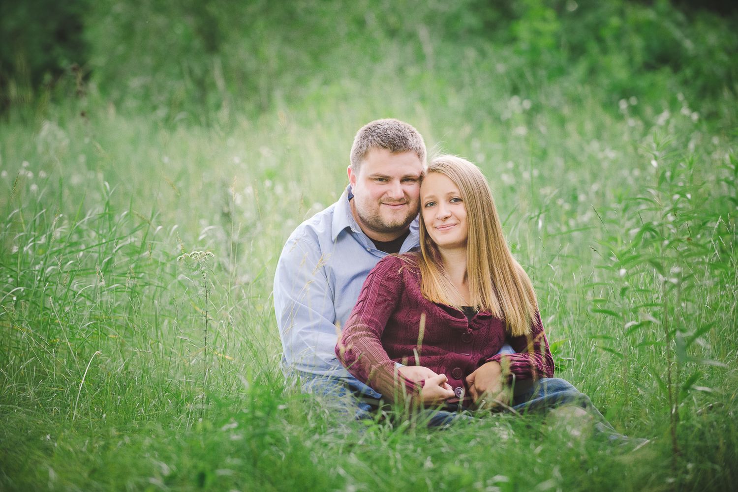 A couple sits together intimately in a field of tall grass on a sunny day.