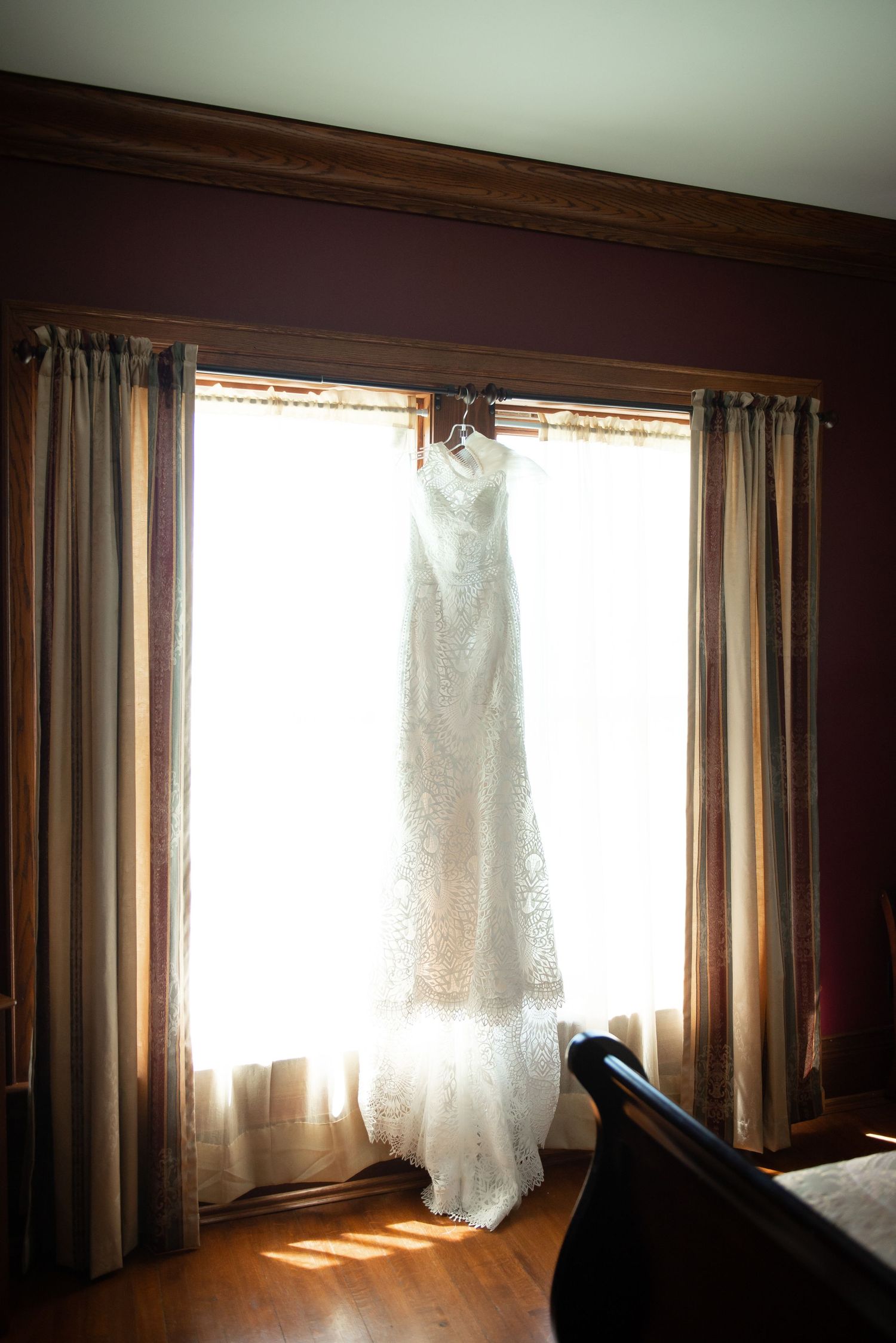 A white wedding dress hangs by a bright window with sunlight streaming through sheer curtains.