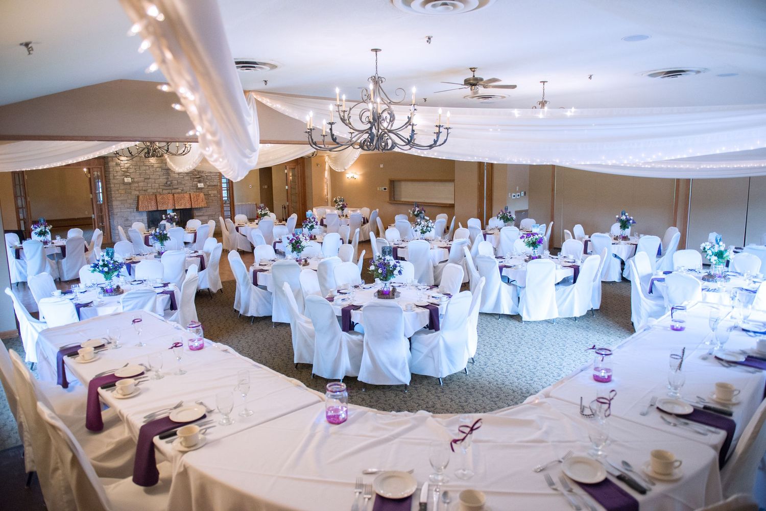 Large wedding reception ballroom with white-covered chairs and tables decorated with purple accents and soft lighting. Andover wedding at Ma