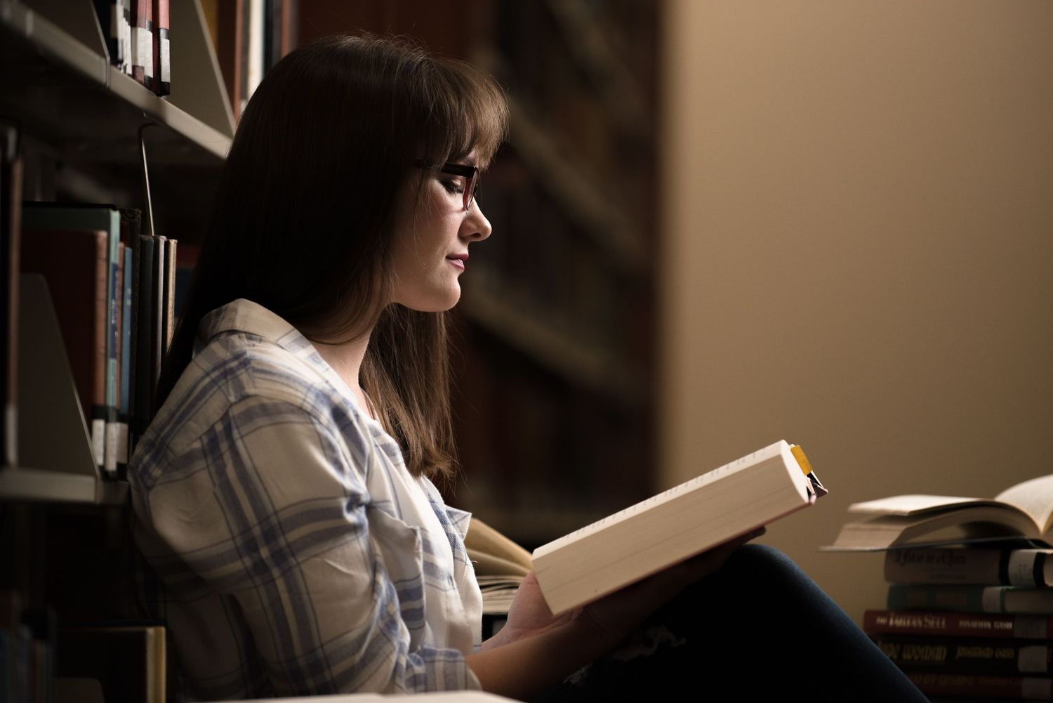 Senior Portrait at St Cloud State in white shirt reads a book near window while sitting in library with warm lighting and bookshelves.