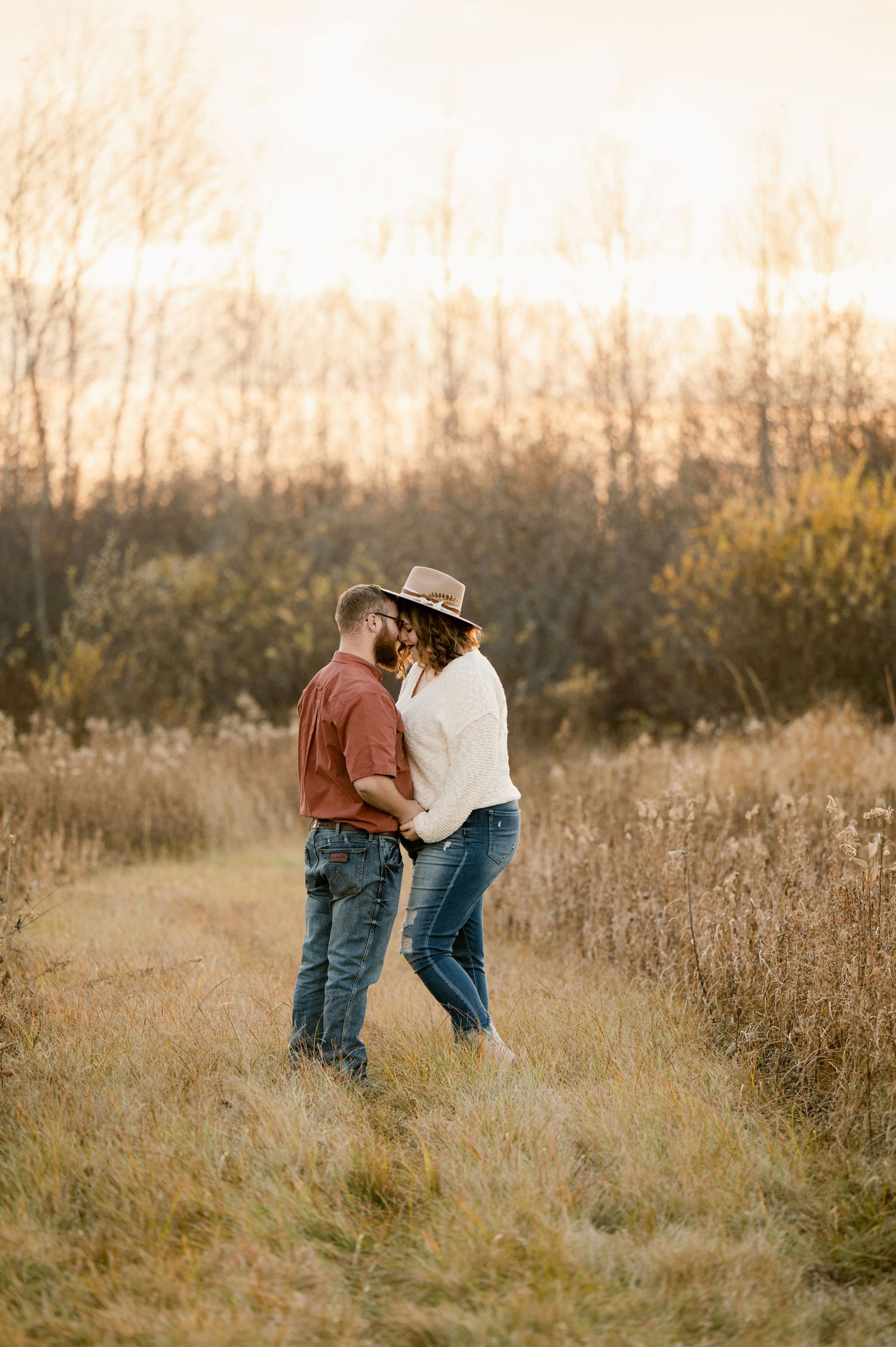 A romantic couple embraces in a golden field at sunset with autumn trees in the background. St cloud area engagement photos.