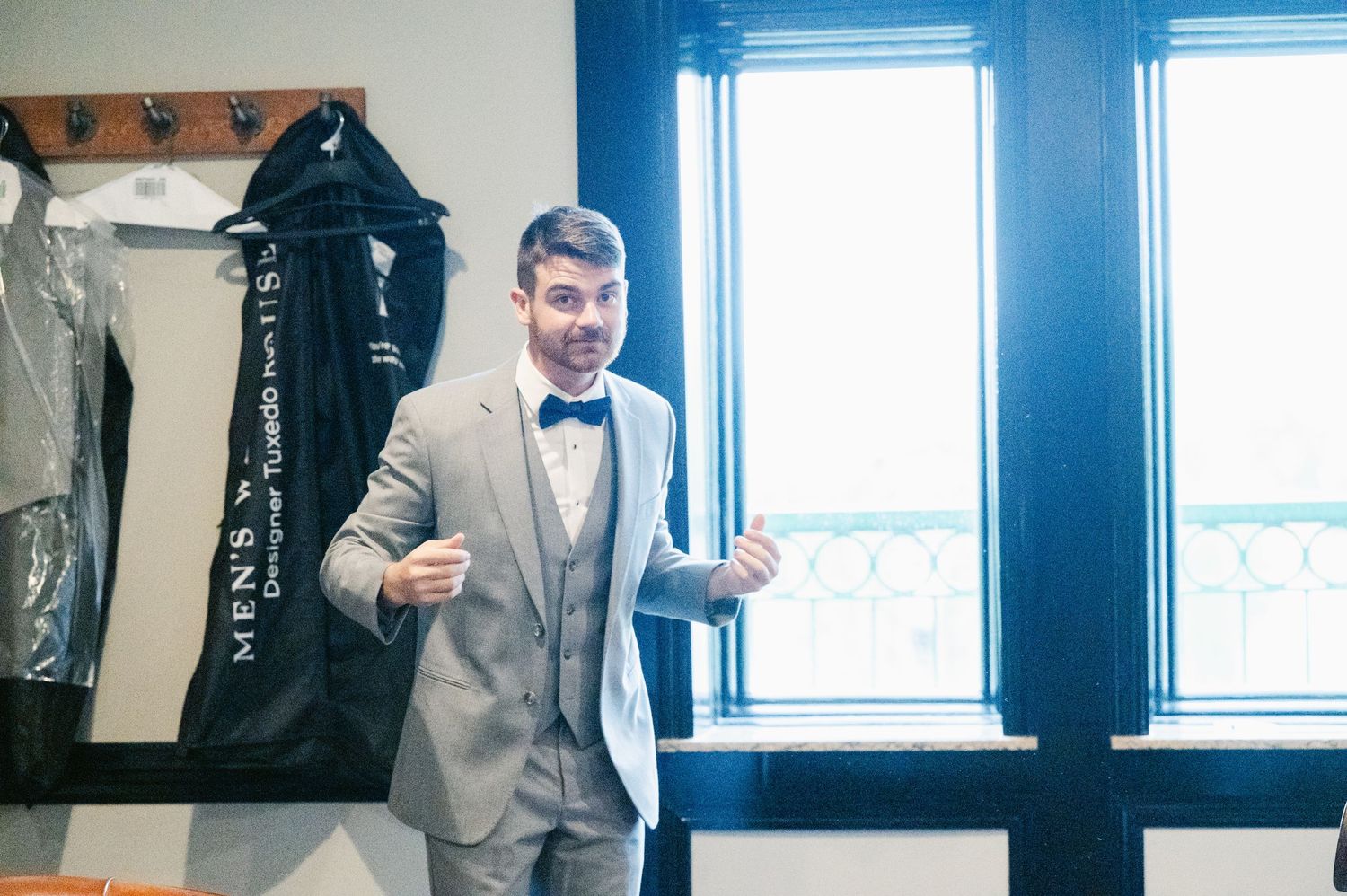 A person adjusts their classic bow tie while standing by a bright window in a light colored suit.