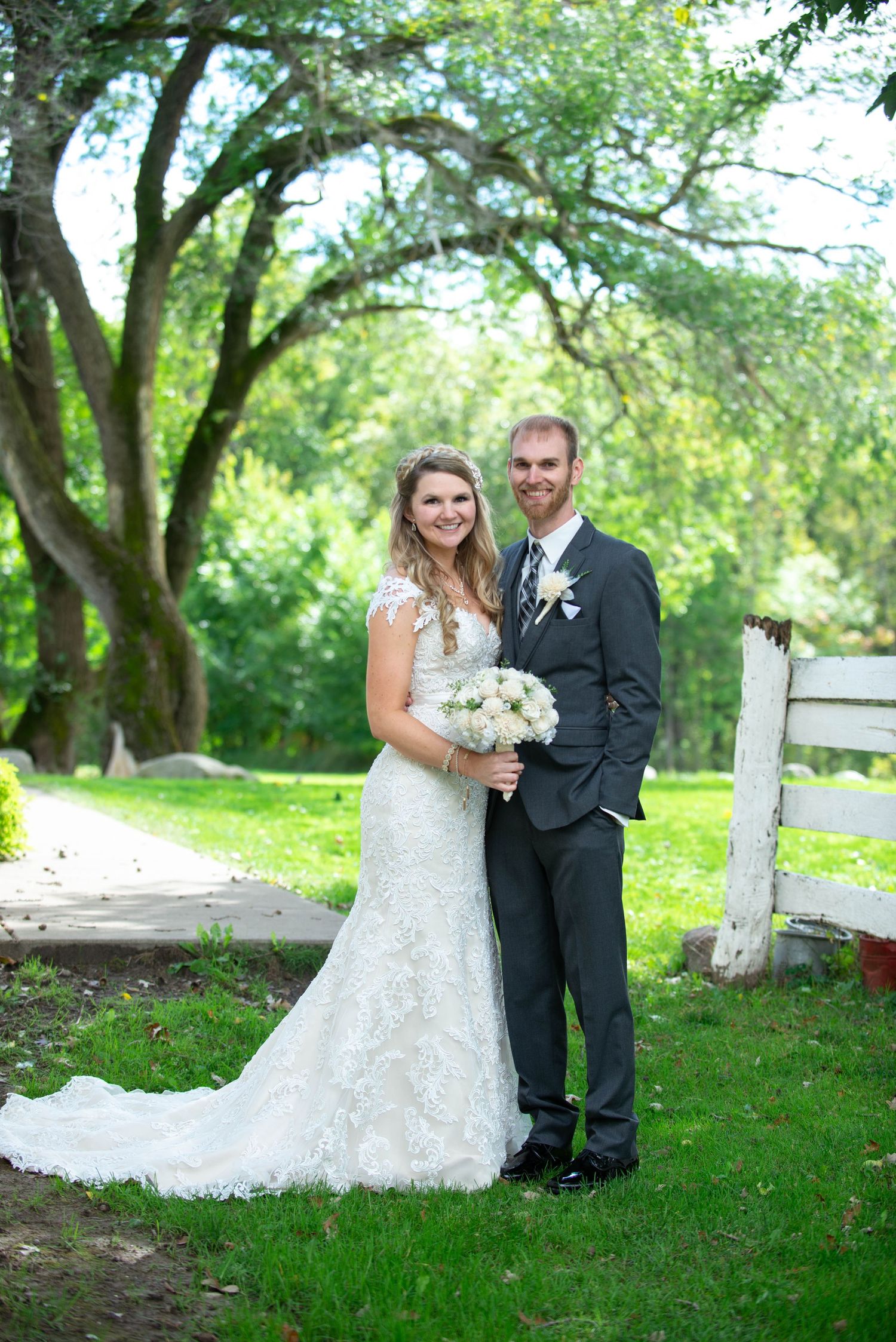 A wedding photo of a couple in formal attire standing on grass with a white fence and trees in the background. Rum River Barn Milaca Minneso
