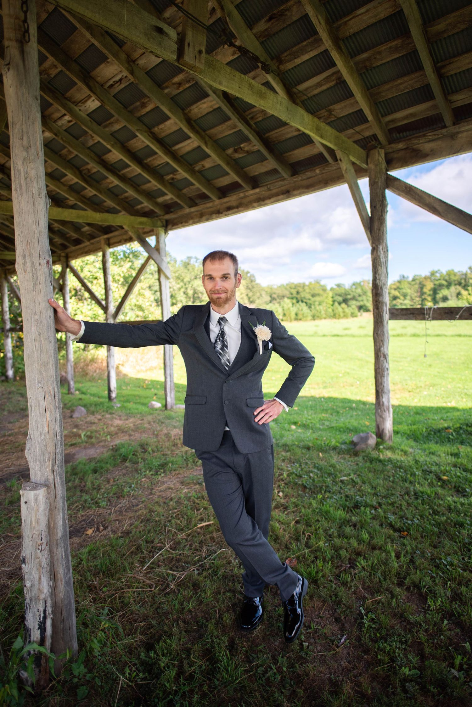 A groom in a dark suit poses casually under a rustic wooden pavilion with a pastoral field in the background.