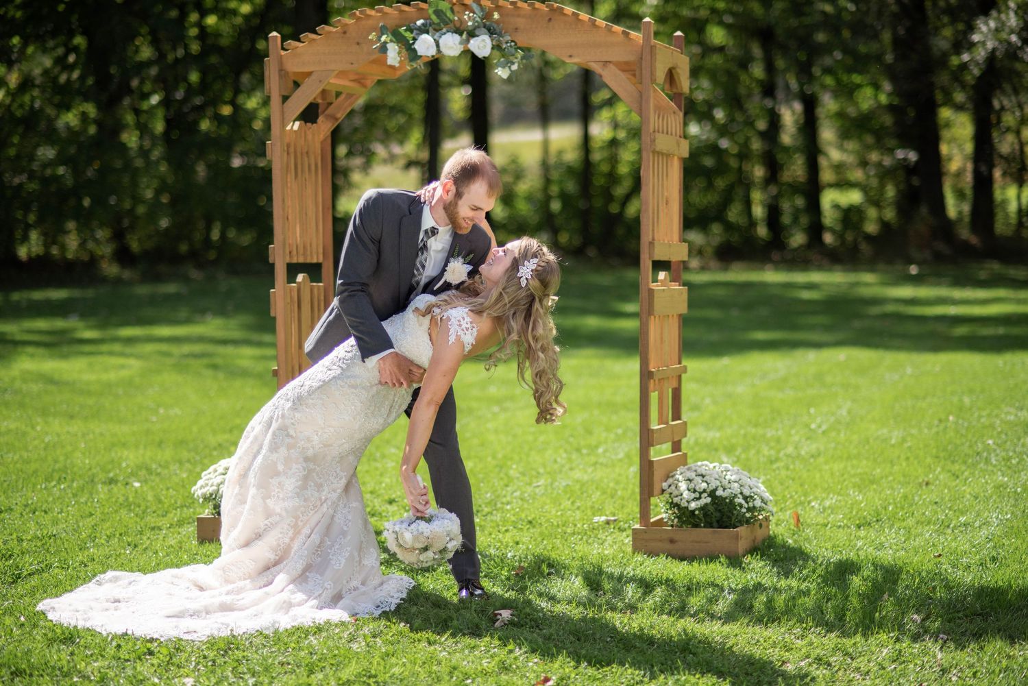 Romantic dip kiss between newlyweds under a wooden wedding arch decorated with white flowers in a garden setting.