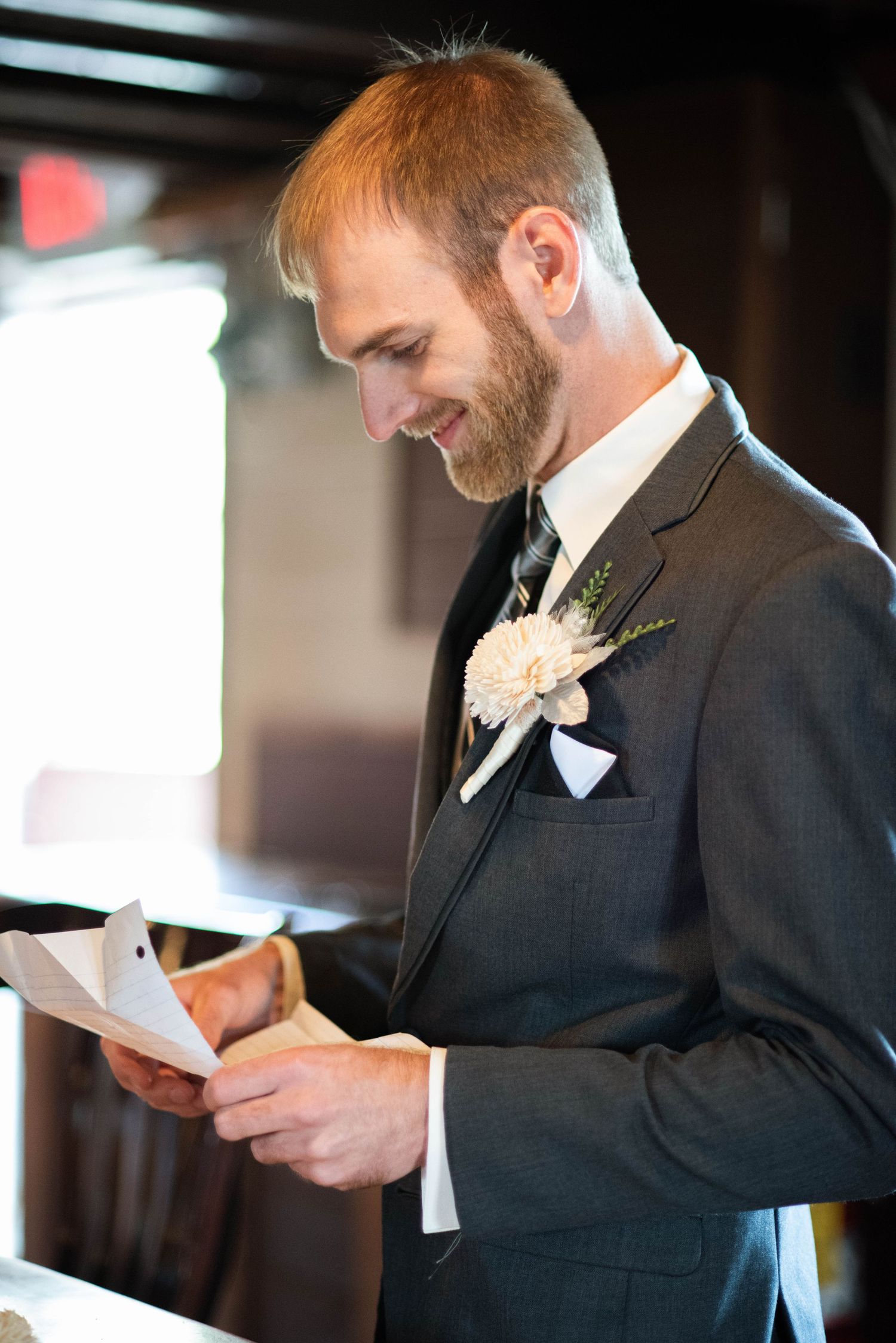 A groom in a dark suit with a light boutonniere reads from a card during a wedding ceremony.