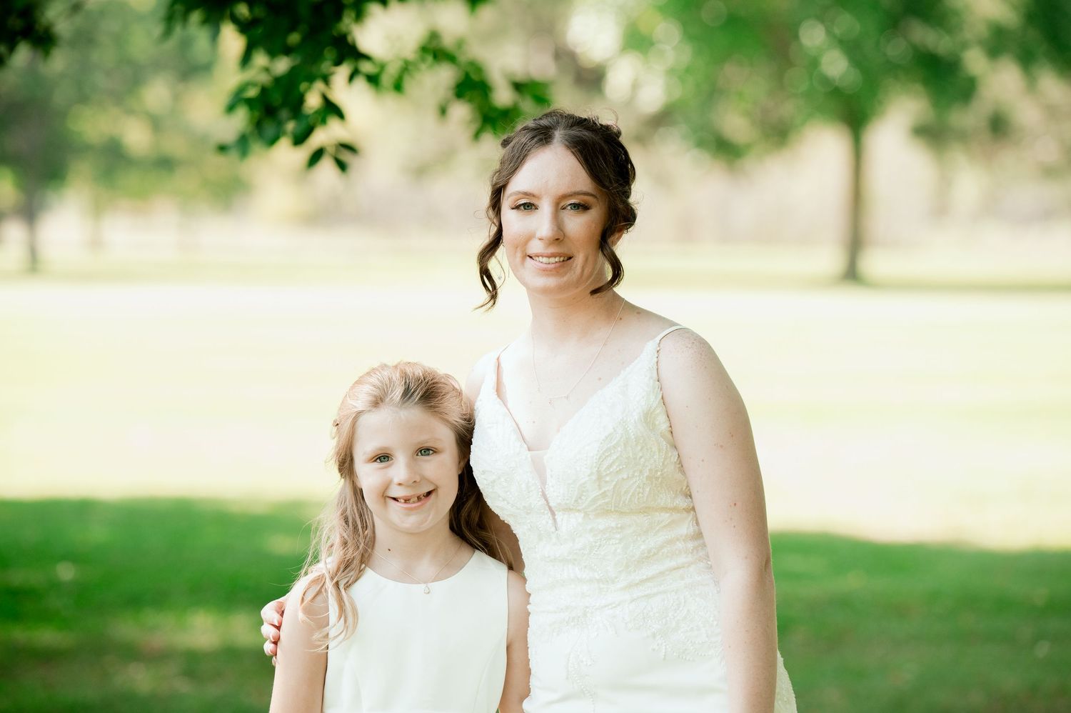 Bride in a white lace dress poses outdoors in a sunny park setting with soft natural lighting.