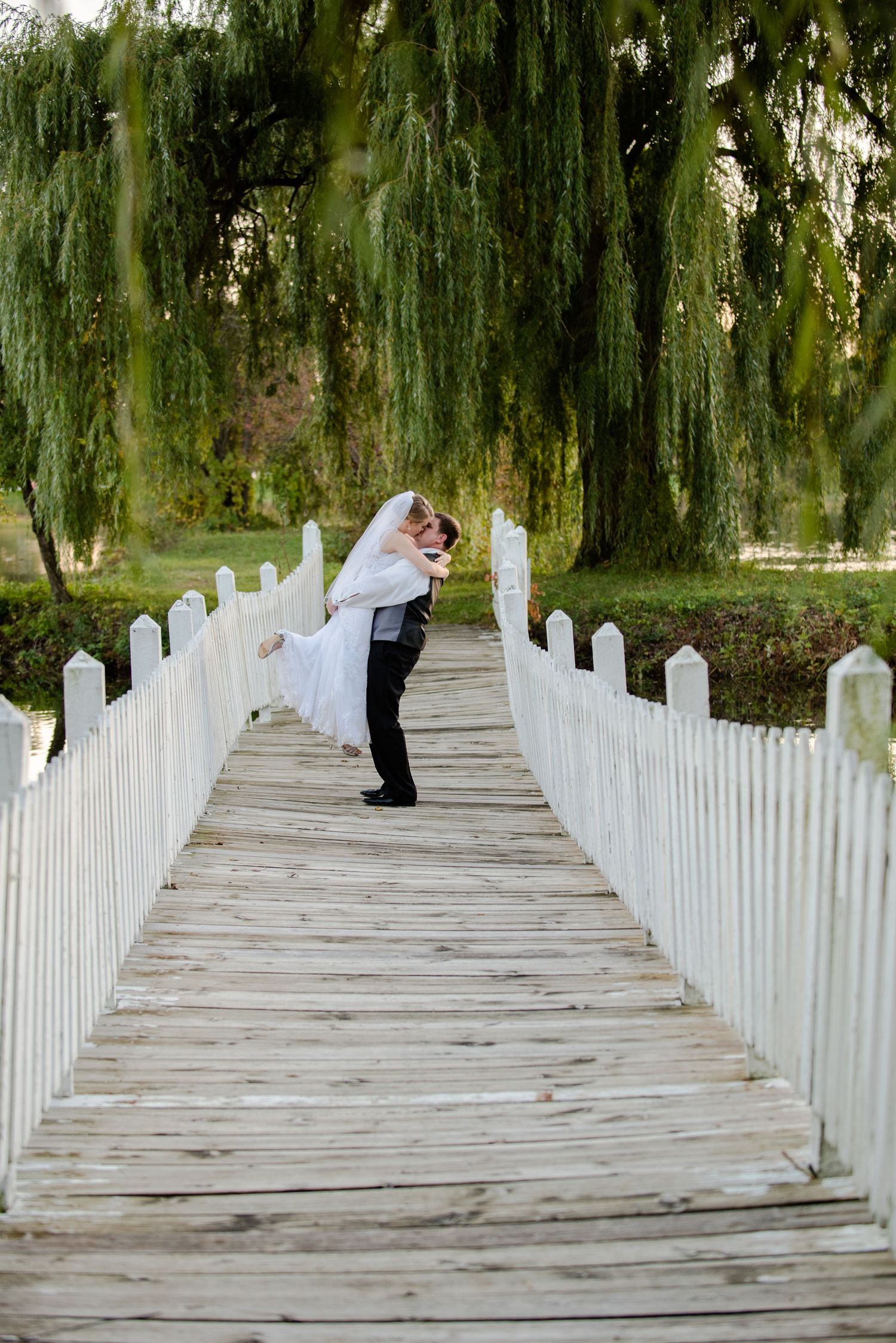 A couple shares a romantic lift moment on a white wooden bridge surrounded by cascading willow trees. Andover wedding at Magestic Oaks Golf