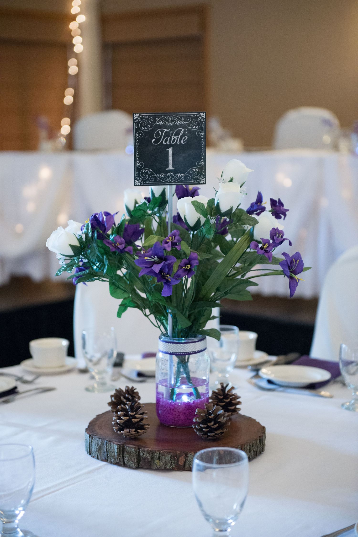 Wedding reception centerpiece with purple and white flowers in mason jar on wood slice with pinecones and chalkboard table number. Andover w