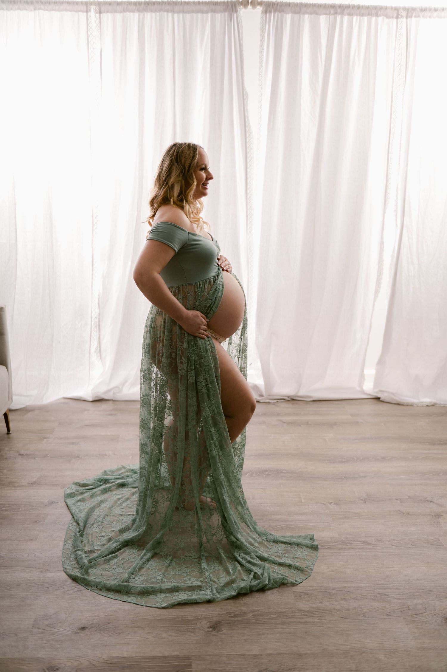 Expectant mother poses in sage green lace maternity gown against white curtain backdrop during intimate photo session.