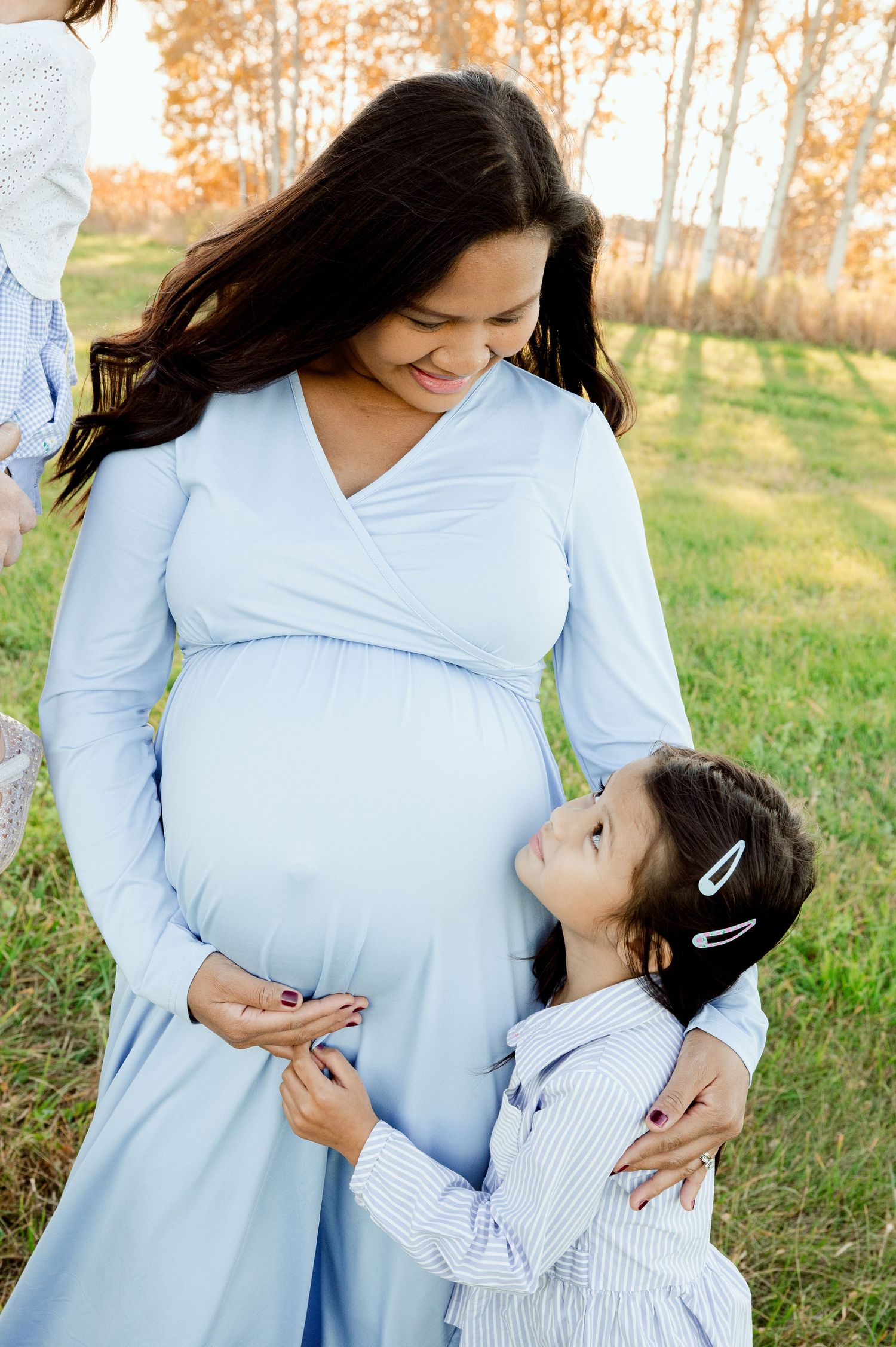 A pregnant woman in a light blue dress shares a tender moment with a young child outdoors.