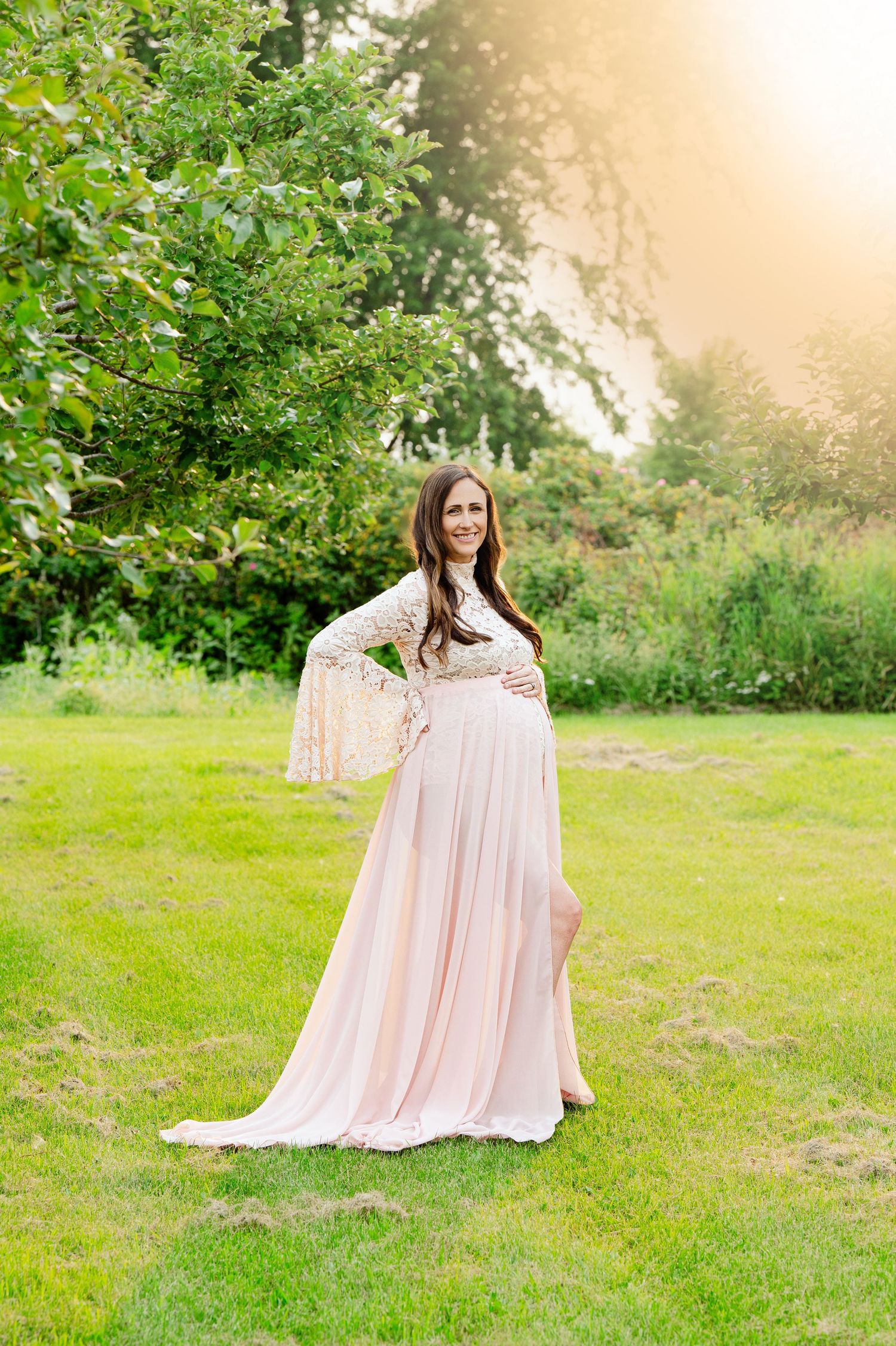 A woman in a flowing pink dress poses in a sunlit garden during a maternity photoshoot.