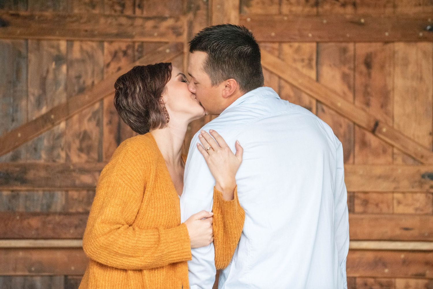 A couple in coordinated yellow and white outfits share a romantic embrace against a rustic wooden barn wall.