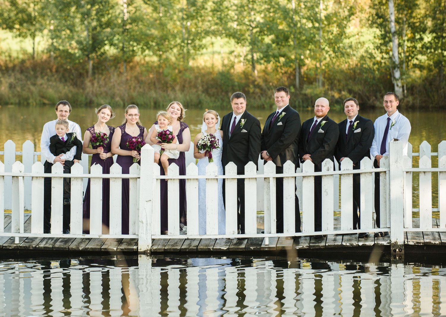 Wedding party poses on wooden dock with white fence overlooking reflective water at sunset. Andover wedding at Magestic Oaks Golf Club