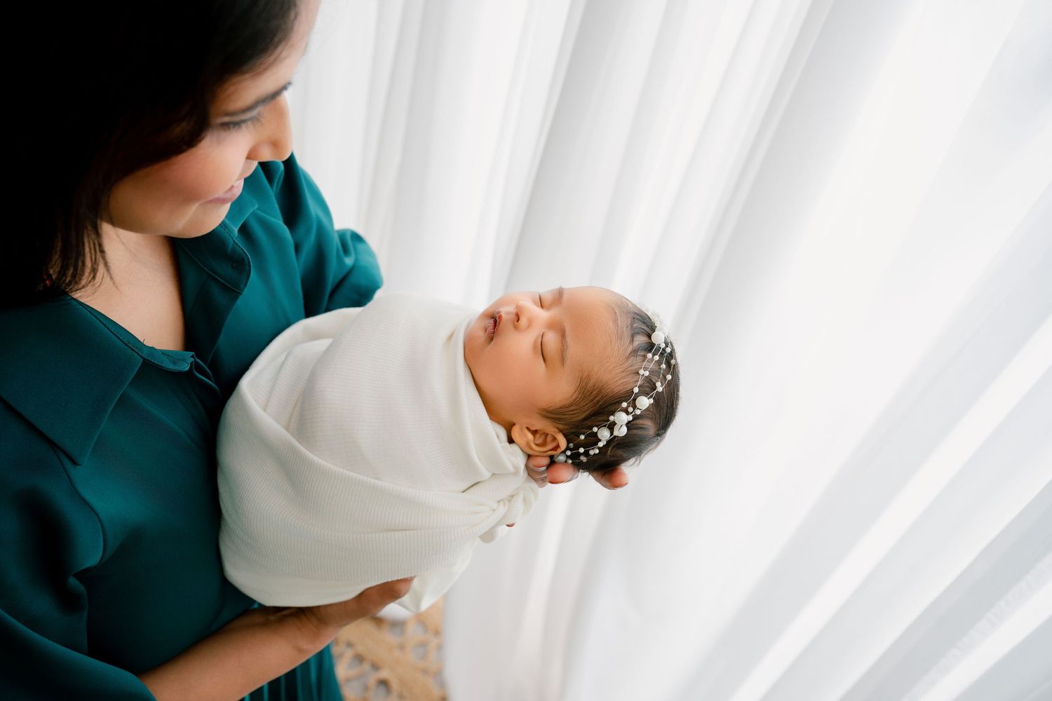 Mother in emerald dress cradles swaddled newborn against white backdrop.
