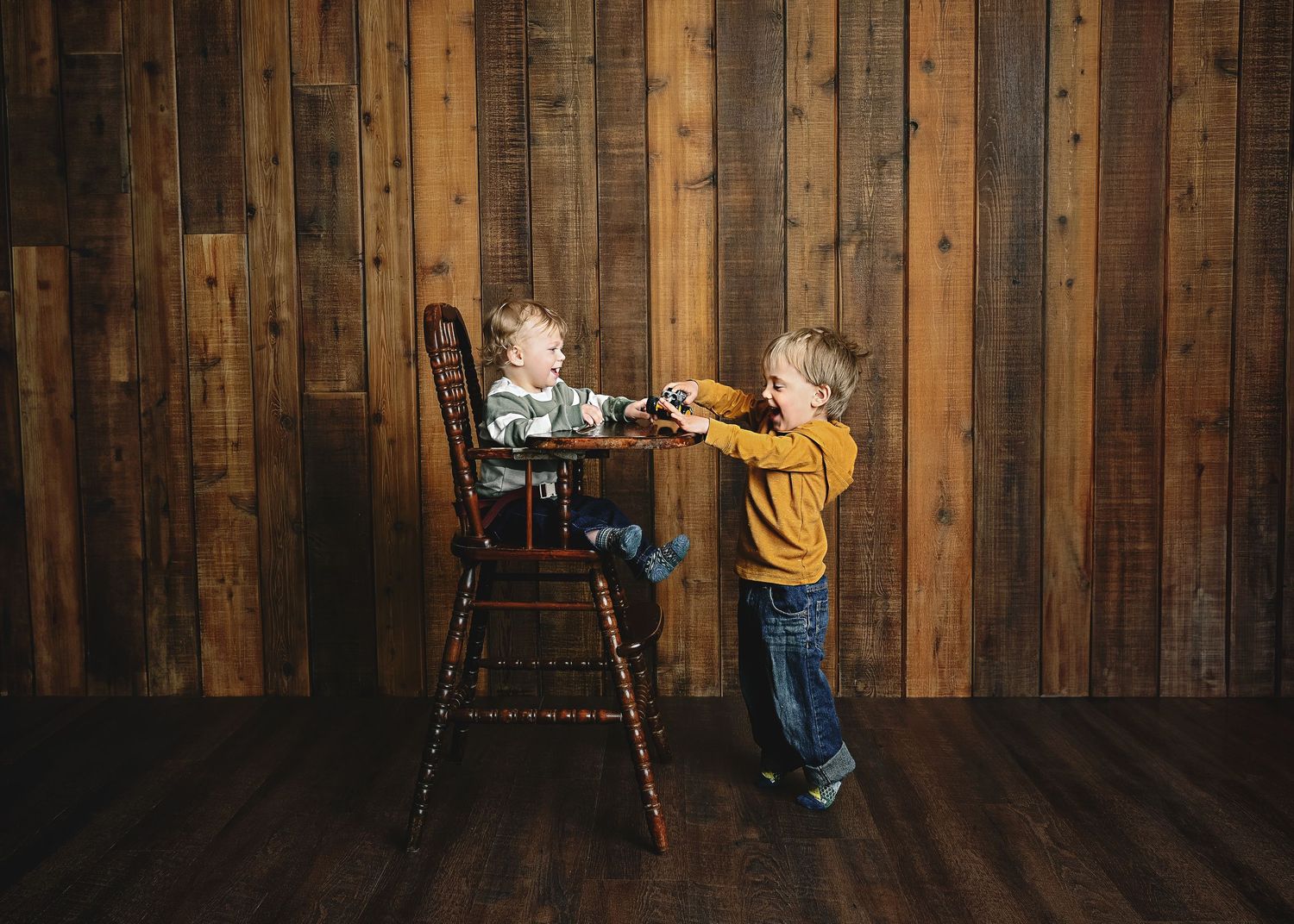 Two young ones interact at wooden high chair against rustic wood panel wall.