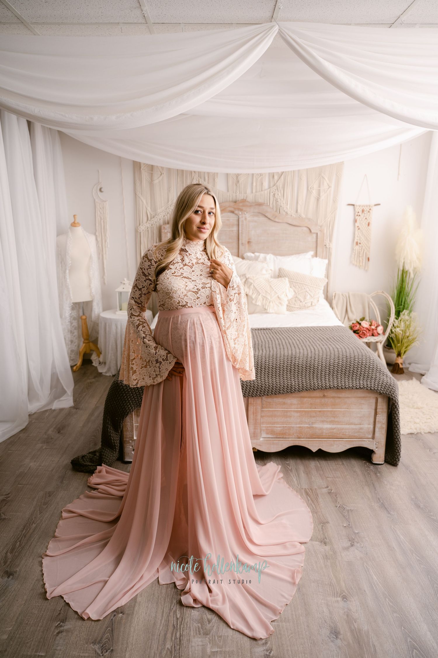A flowing pink maternity gown with lace sleeves is showcased in a romantic bedroom setting with draped white fabric decor.