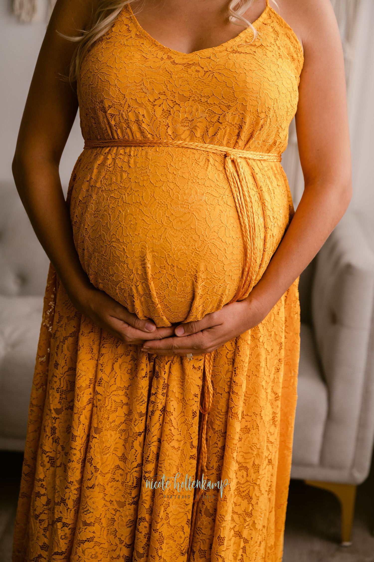 Closeup of pregnant belly in a flowing marigold yellow lace maternity dress showing gentle hands cradling the bump.