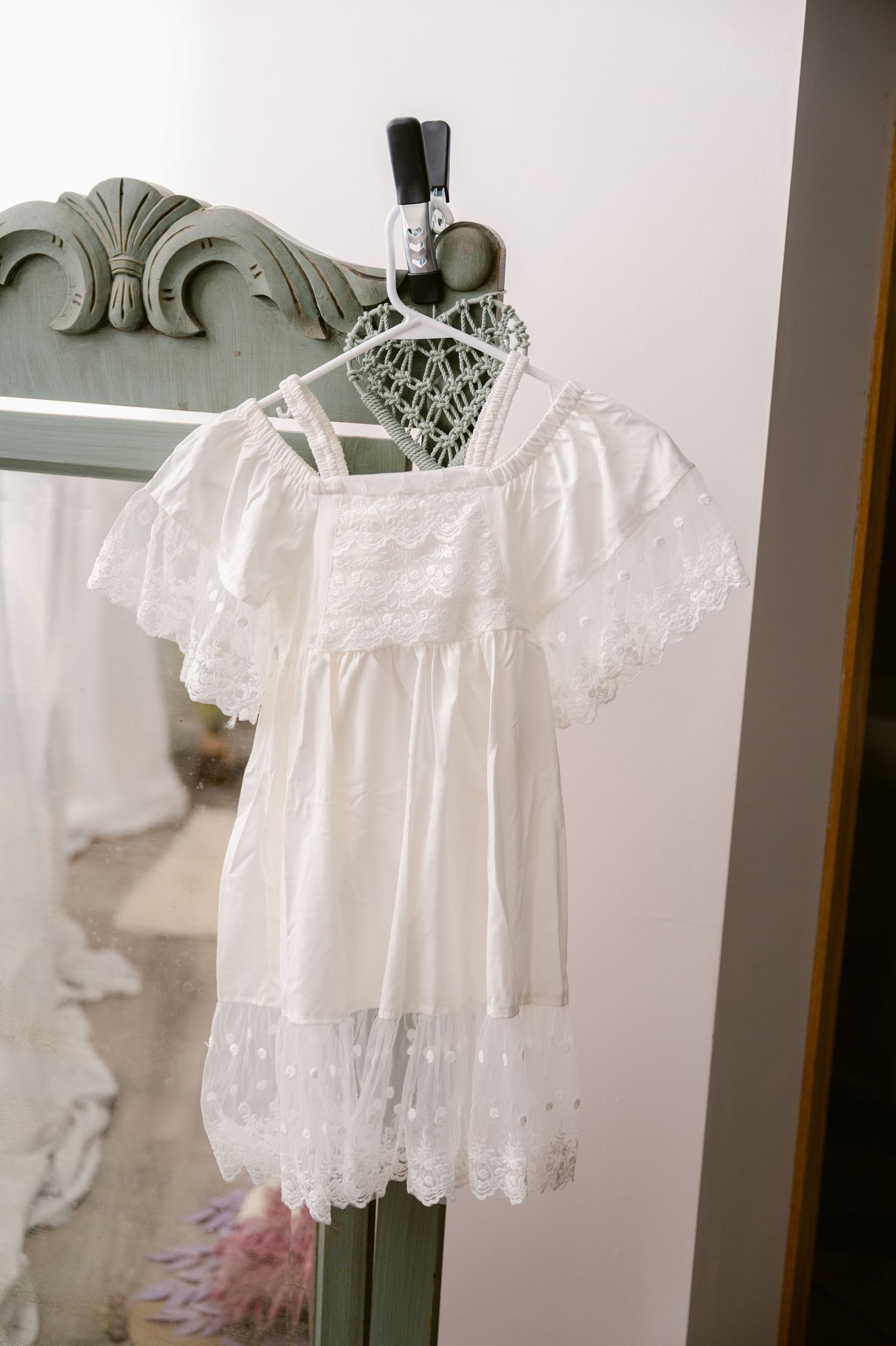 A delicate white lace baby dress with puff sleeves hangs on a decorative wooden hanger against a light background.