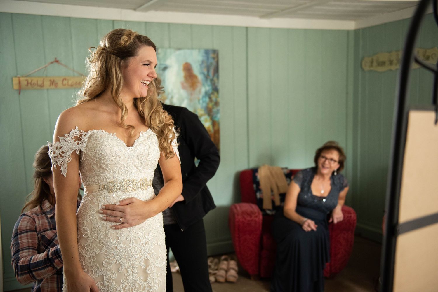 A bride in a lace wedding dress stands in a mint green room during preparations.