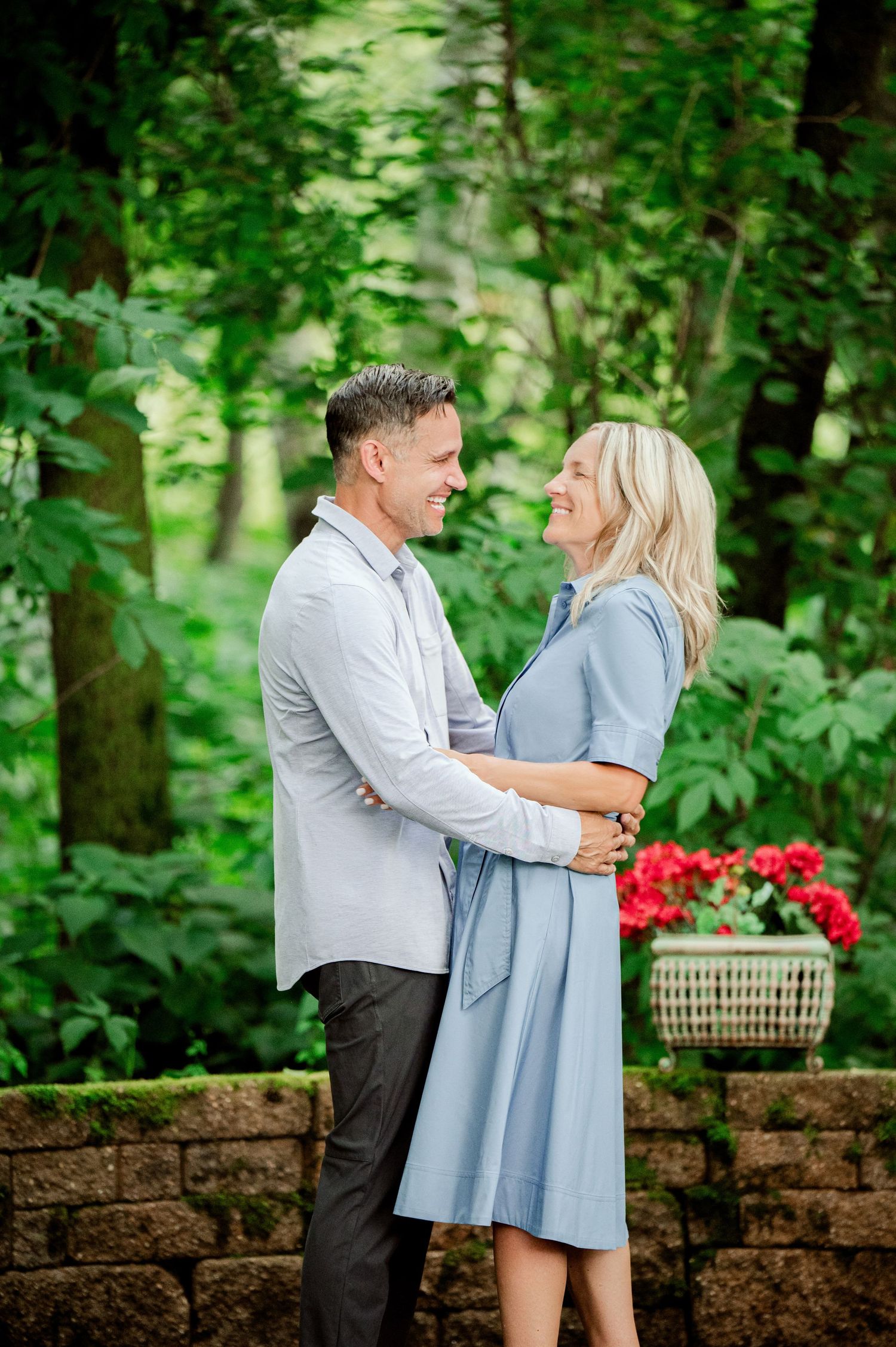 Clear Lake MN Family Photos: Couple shares an intimate moment on a garden patio with red flowers and green foliage behind them.