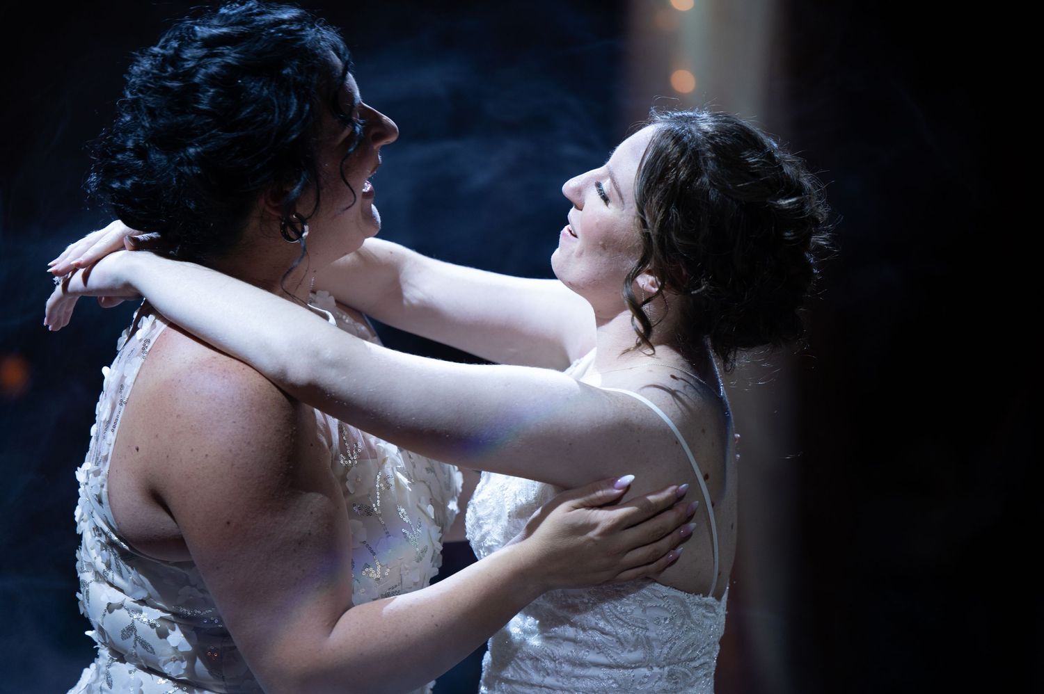Two people in wedding dresses share an intimate dance moment in dramatic lighting.
