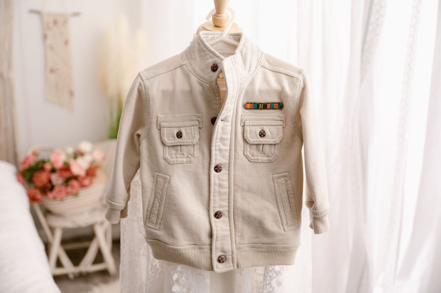 Beige denim jacket for kids with button front and chest pockets displayed against soft white curtain background.
