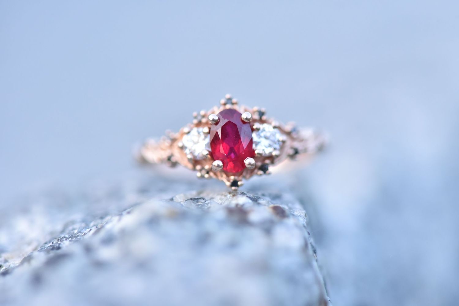 Elegant rose gold ring with oval ruby center stone and small diamond accents photographed on textured blue surface.