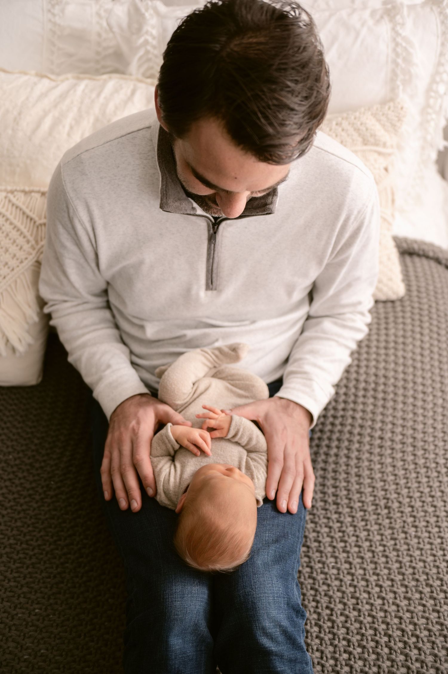 A person in a light colored sweater tenderly holds a small infant on their lap while sitting on a gray couch.