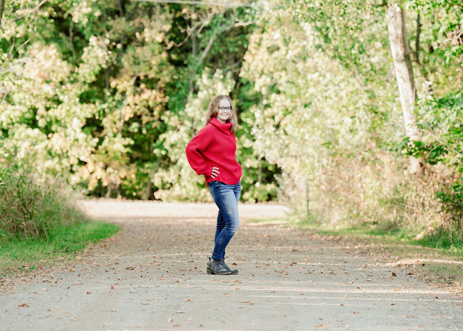 Senior Portrait in Foreston MN in a red sweater and jeans stands on a winding path surrounded by autumn foliage and trees.