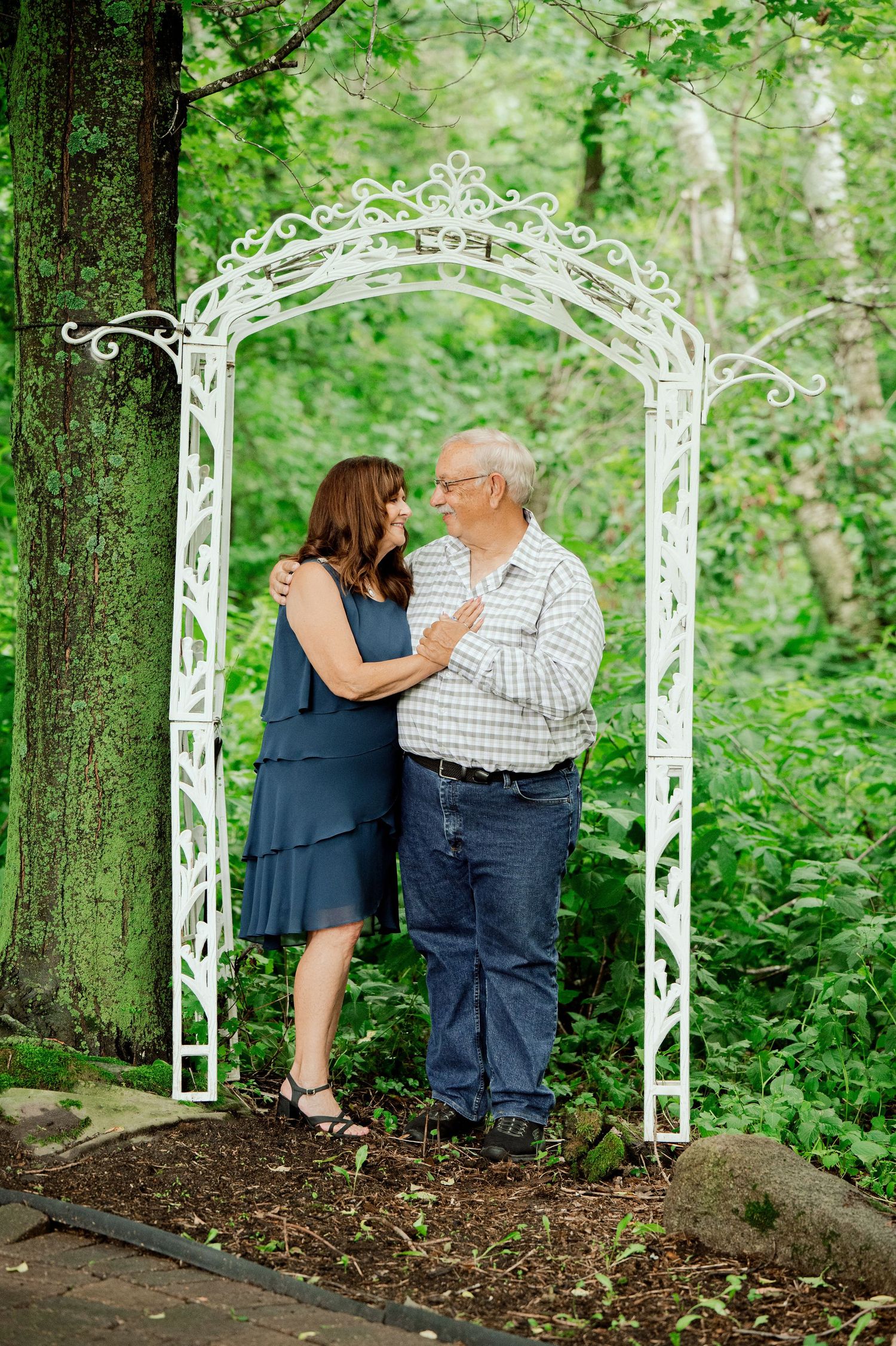Clear Lake Family Couple shares a tender moment under an ornate white garden arch surrounded by vibrant green foliage in a wooded setting.
