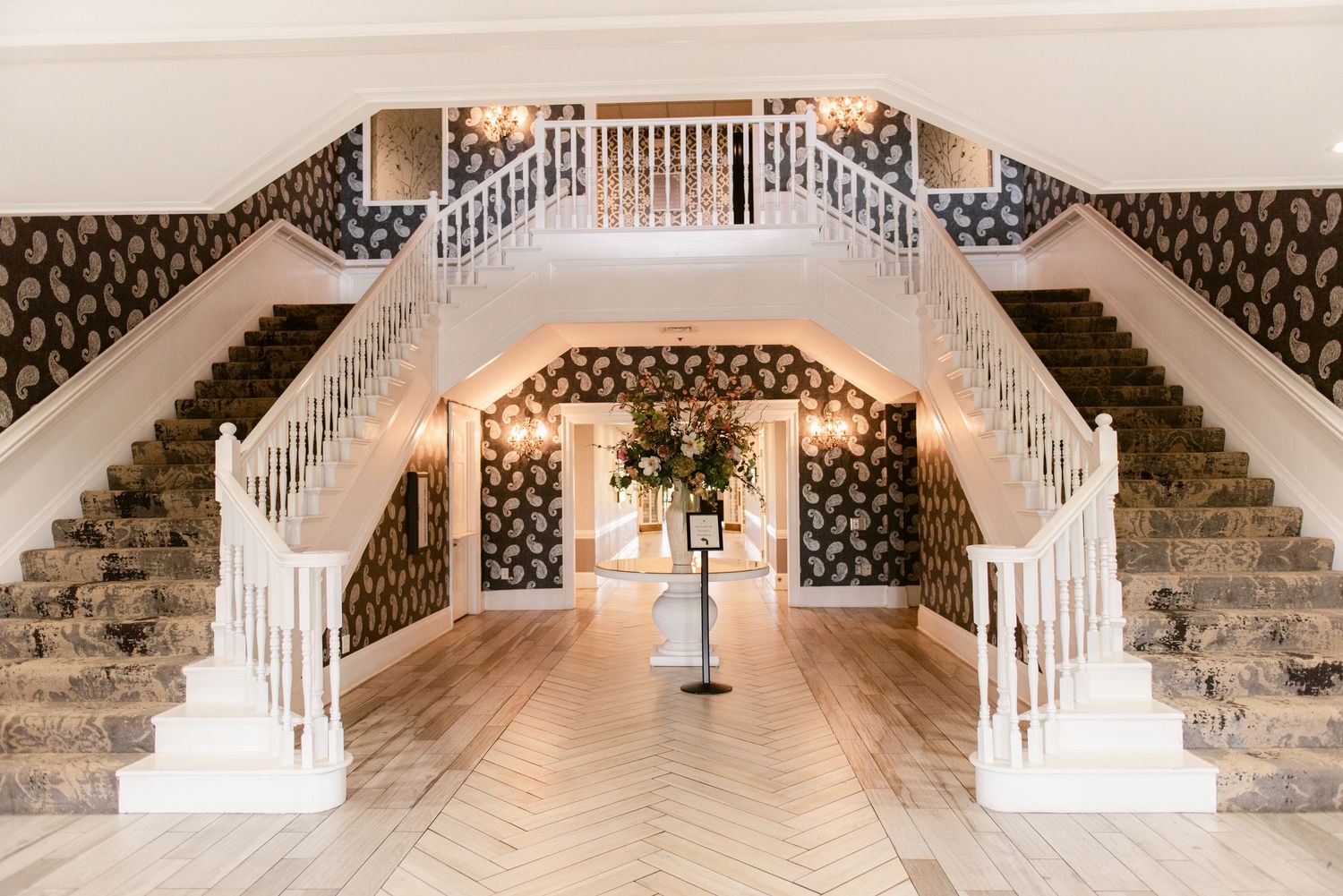 Elegant double staircase with white railings and herringbone wood floor in grand wedding venue entrance.