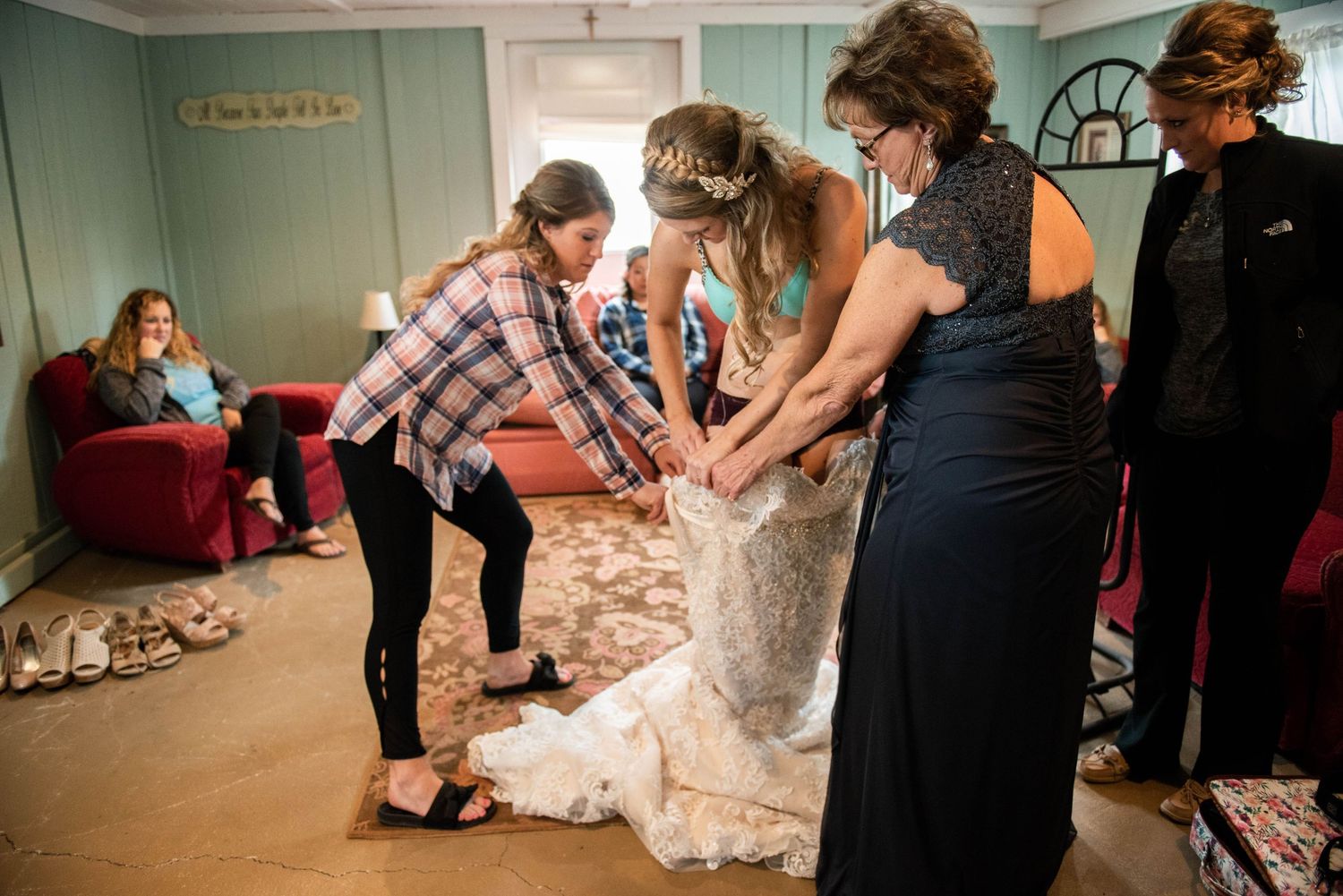 People help adjust the hem of a white wedding dress in a mint-colored room.