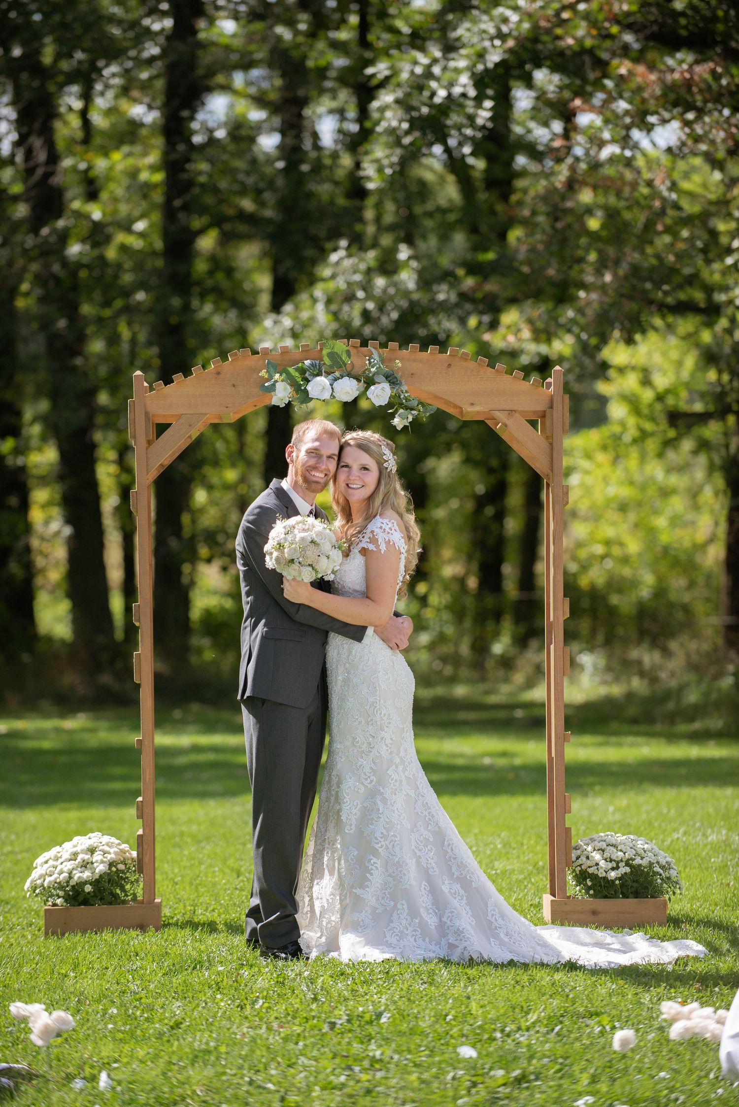 Wedding couple poses under a wooden arch decorated with flowers on a sunny lawn. Rum River Barn Milaca Minnesota Weddings