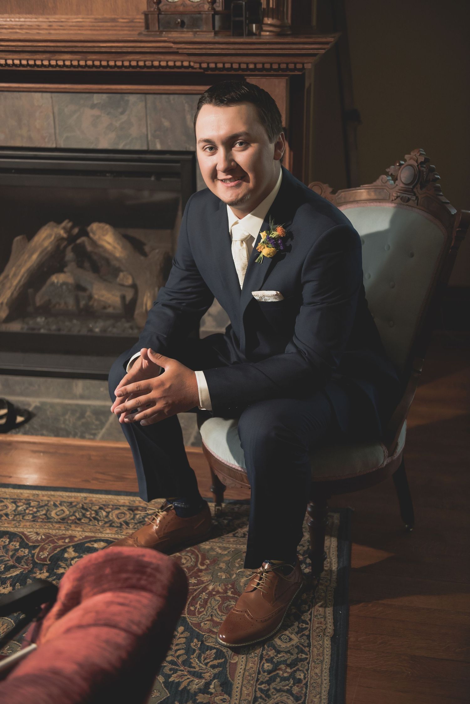 A groom in a navy suit sits in a vintage chair by a fireplace on his wedding day.