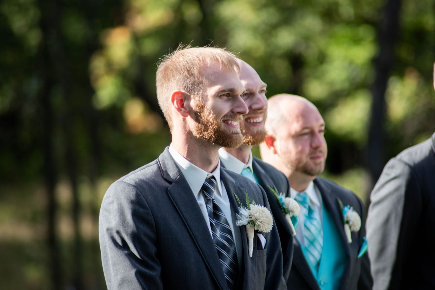 Wedding party members in formal attire smile during an outdoor ceremony with natural greenery in background.