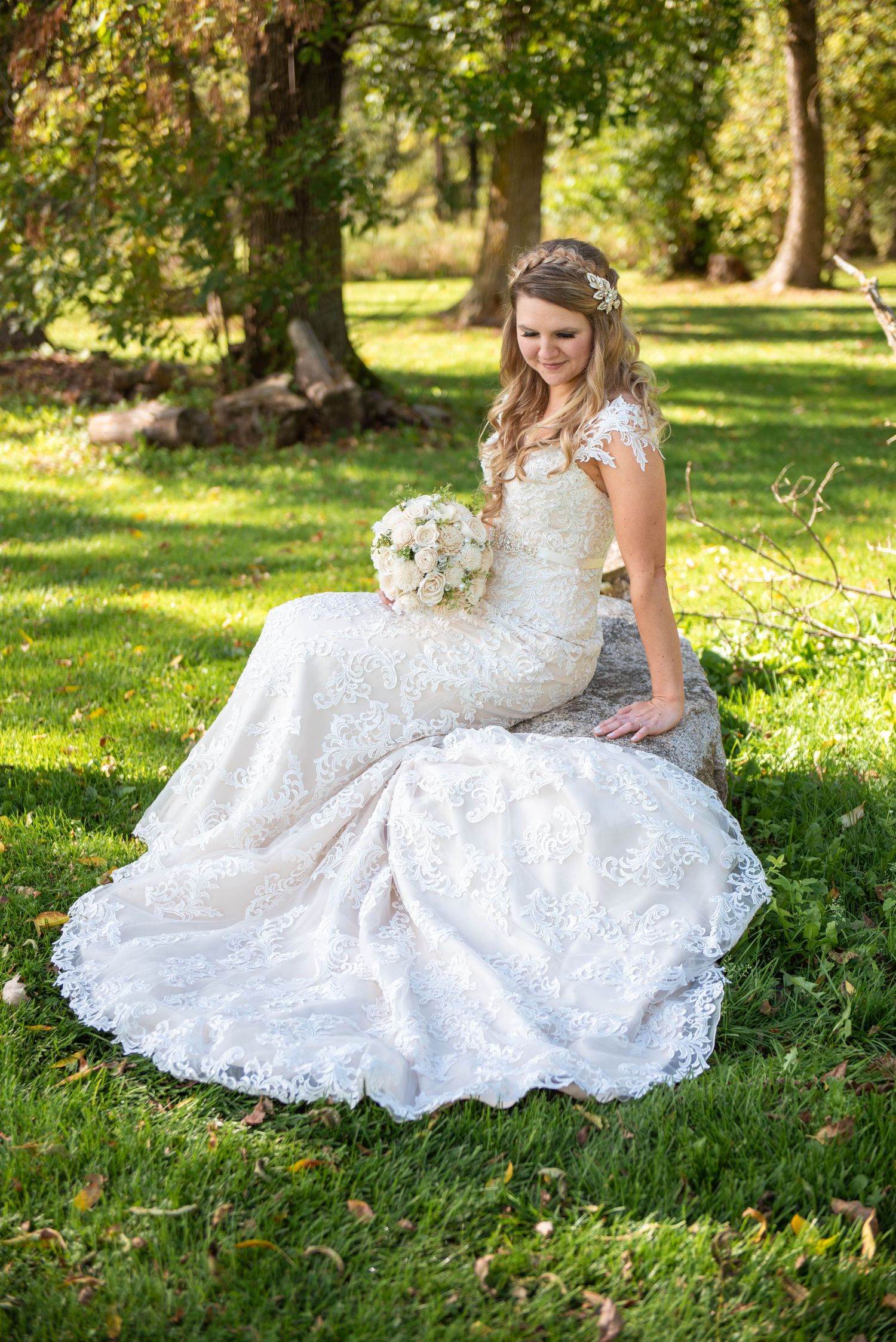 A bride in an elegant lace wedding dress sits gracefully on grass in a sunlit garden setting. Rum River Barn Milaca Minnesota Weddings