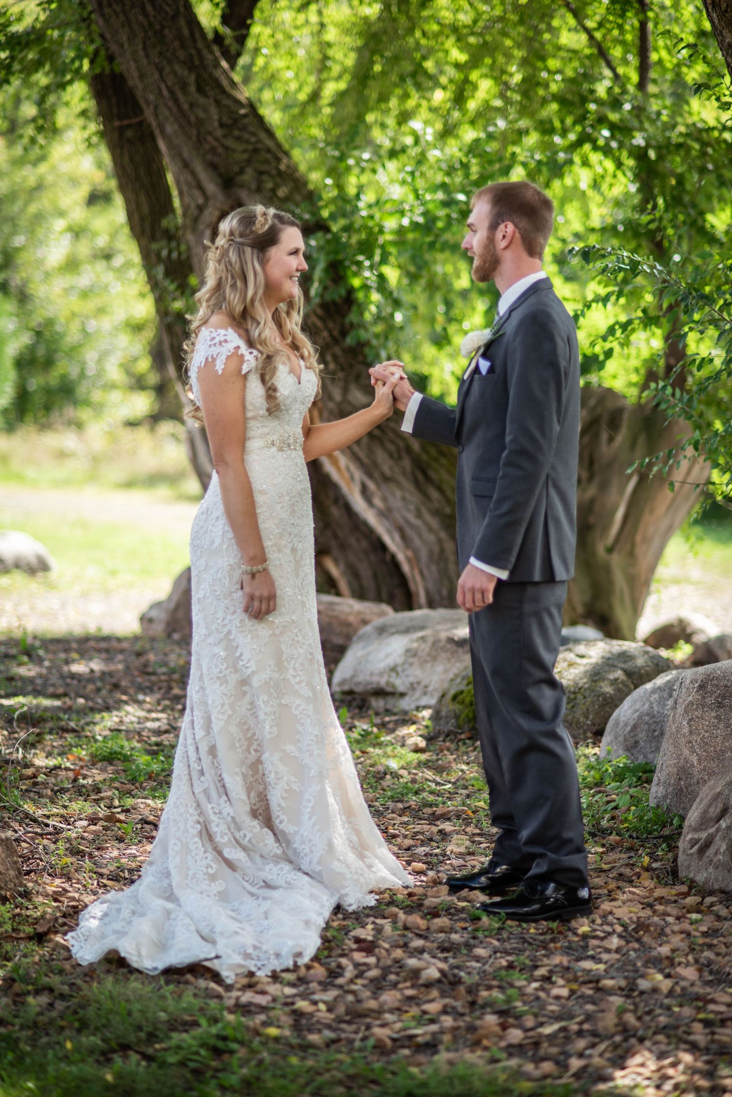 A couple in wedding attire holding hands and dancing under lush green trees in a romantic outdoor setting.