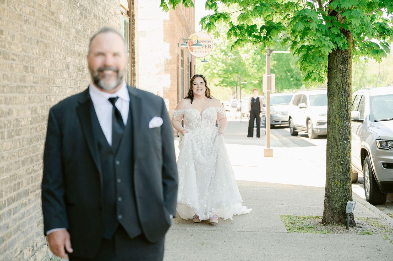 A couple during their first look moment on a sidewalk with green trees and parked cars in the background.
