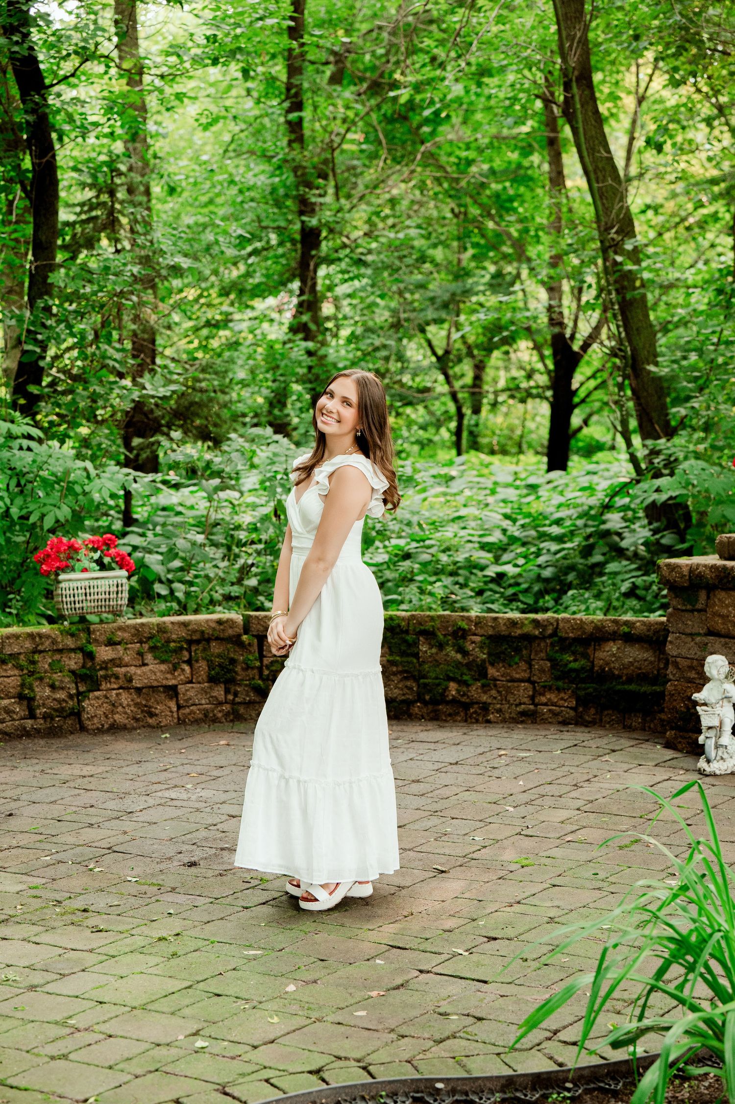 Senior Portrait in Central MN Woman in white summer dress poses on brick patio surrounded by lush green forest garden.