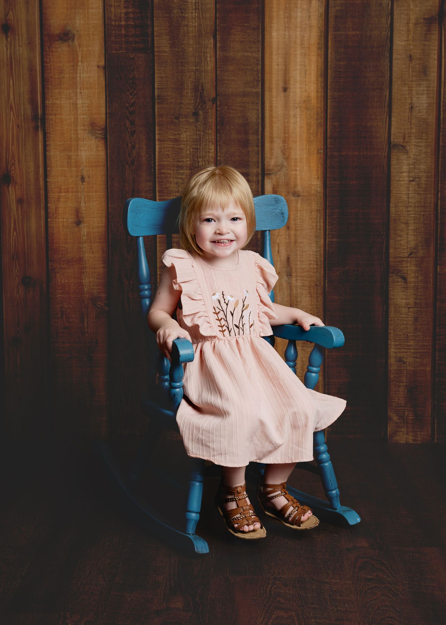 Young child in pink dress sitting on blue rocking chair against wooden wall.