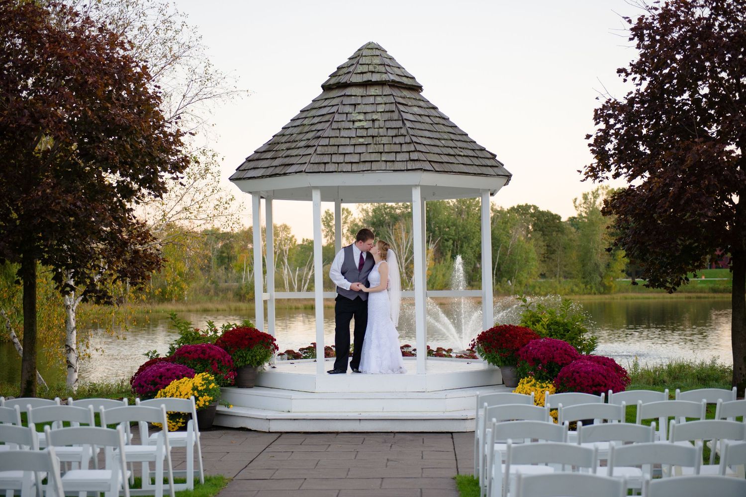 Wedding ceremony setup with a white gazebo overlooking a pond and red flower decorations at sunset. Andover wedding at Magestic Oaks Golf Cl